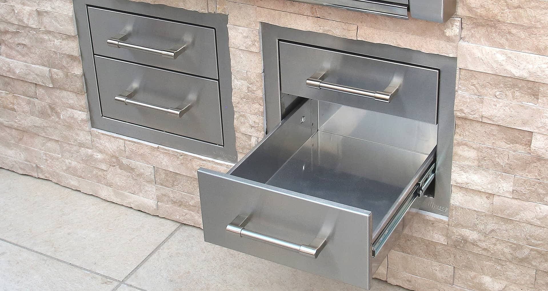 Dos cajoneras de acero inoxidable empotradas en un muro de piedra clara: una con dos cajones cerrados y otra con el cajón inferior abierto sobre sus correderas; piso de loseta clara.