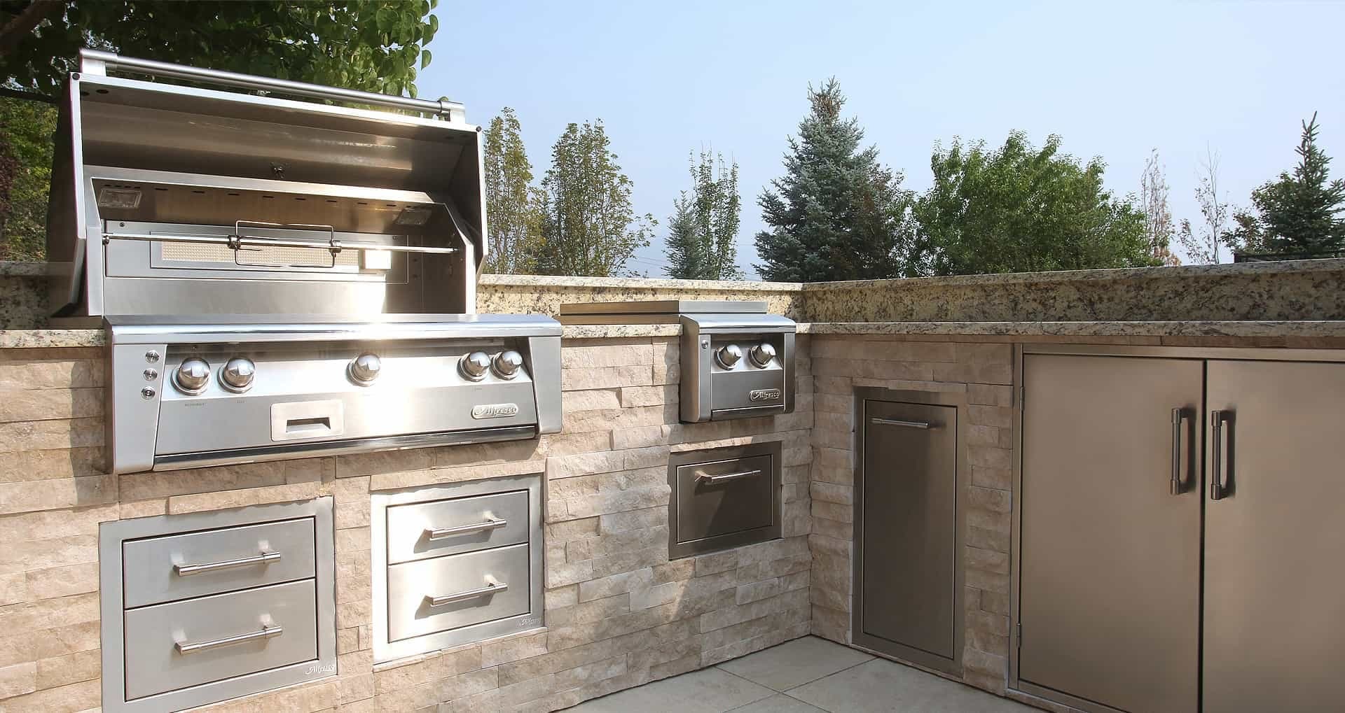 Isla exterior en L de piedra clara con cubierta de granito: asador de acero inoxidable con la tapa abierta y cinco perillas, quemador lateral doble con tapa, dos cajoneras, un cajón, una puerta sencilla y una puerta doble; al fondo, árboles y cielo despejado.