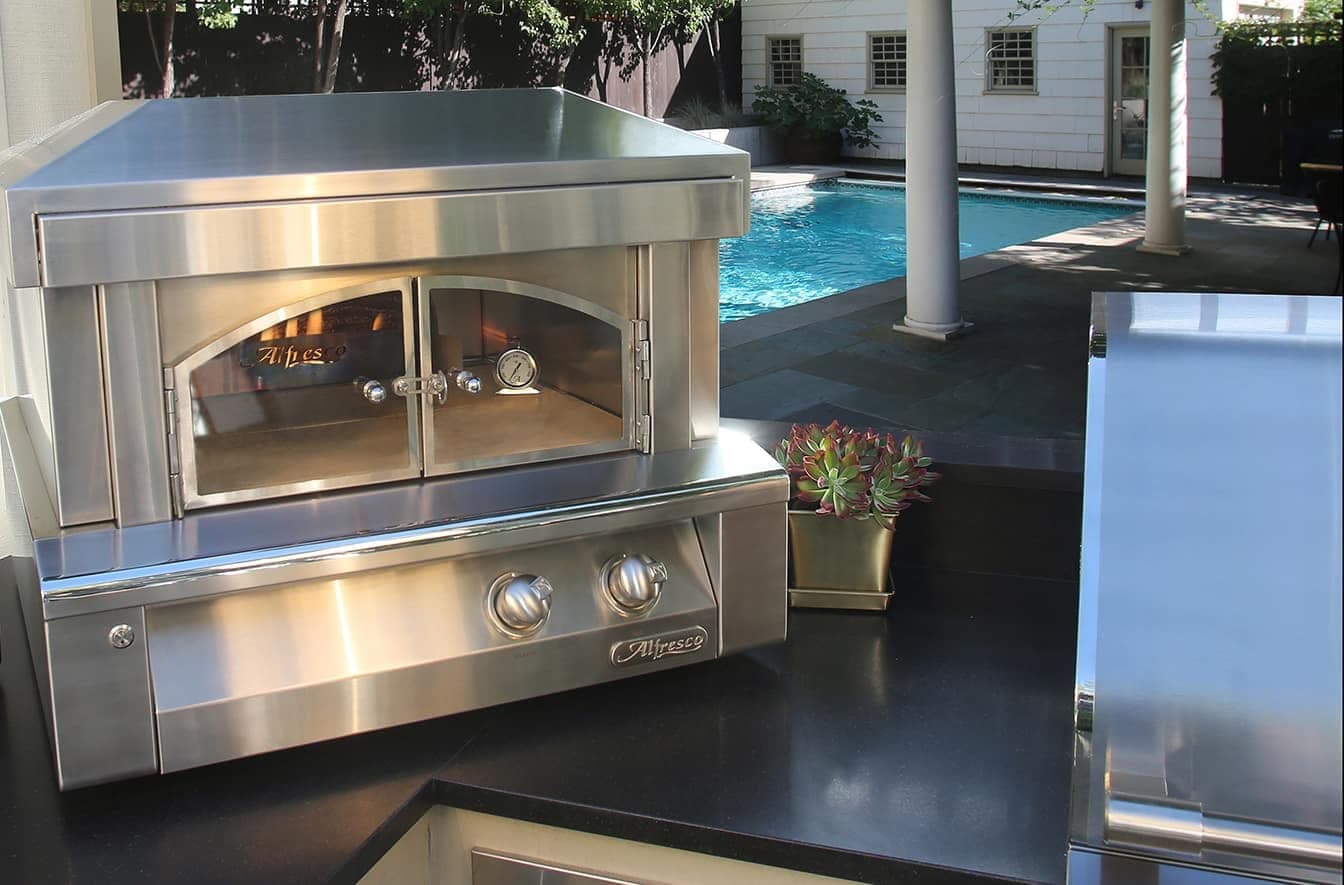 Horno para pizza de acero inoxidable sobre una cubierta negra, con puertas de vidrio en arco, termómetro y dos perillas; al lado, suculentas en una maceta dorada y, al fondo, una alberca, columnas y una casa blanca.