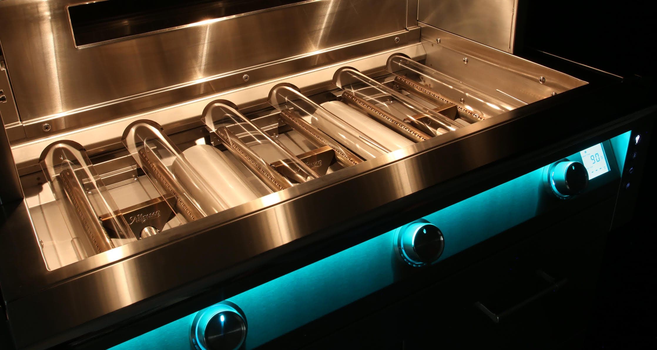 Interior de un asador de acero inoxidable con la tapa abierta, sobre fondo oscuro: cubiertas curvas de vidrio transparente sobre los quemadores, iluminadas con luz cálida, y al frente un tablero con luz turquesa, tres perillas con anillos iluminados y una pantalla digital que marca 90 °F.