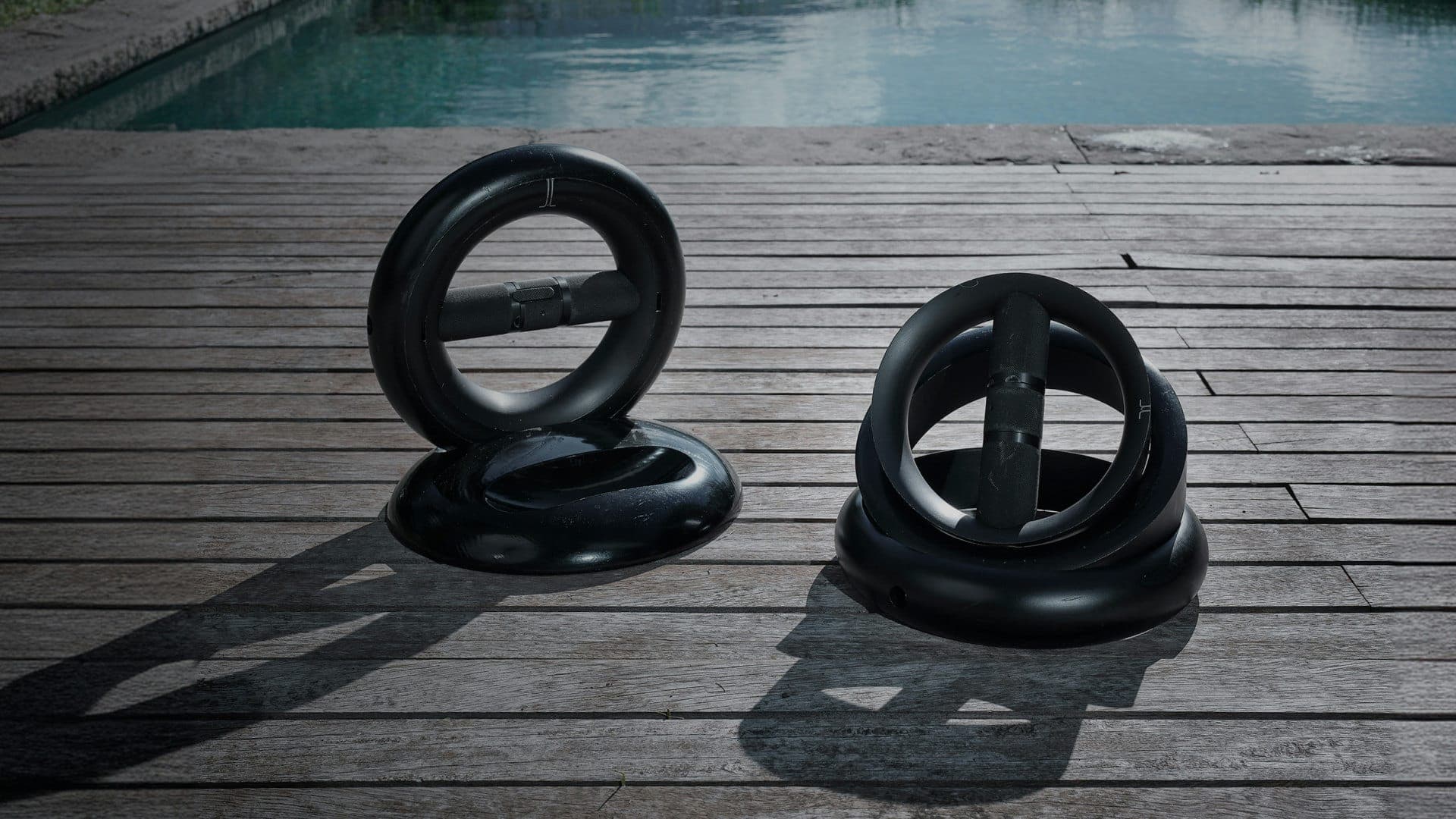 Dos juegos de mancuernas negras de forma circular, cada uno sobre su base, en una terraza de duela gris junto a una alberca.