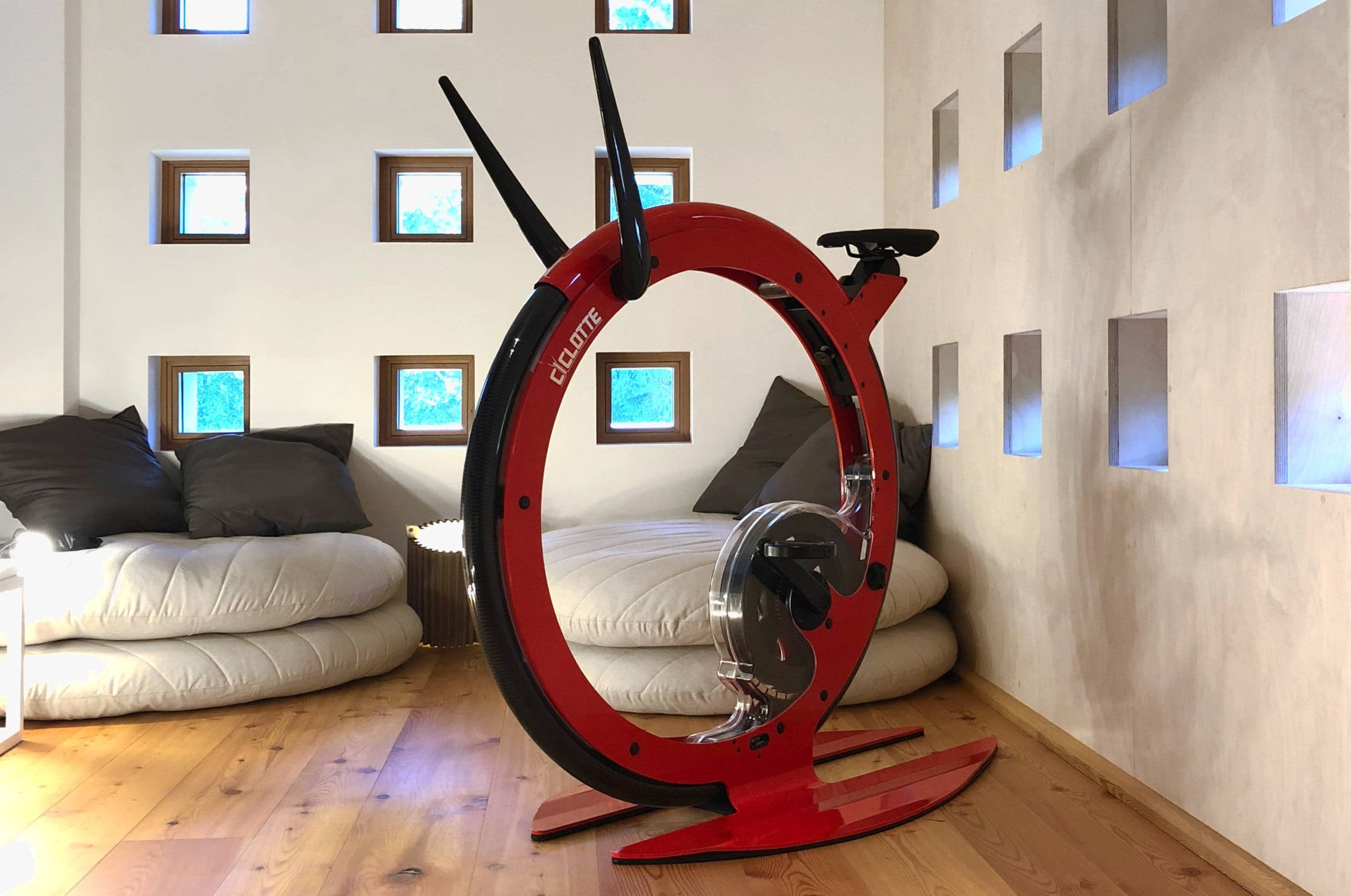Bicicleta fija Ciclotte roja de rueda única en una habitación de muros blancos con ventanas cuadradas pequeñas, futones claros con cojines grises y piso de duela.