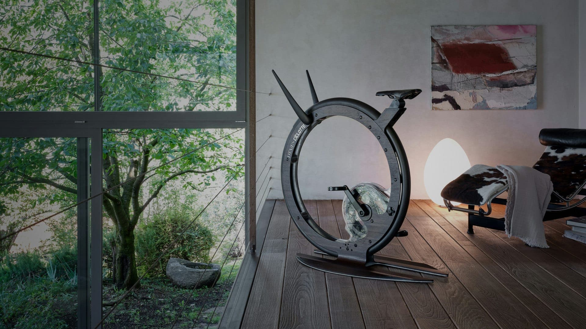Bicicleta fija de rueda única gris oscuro sobre piso de duela, junto a un ventanal con árboles