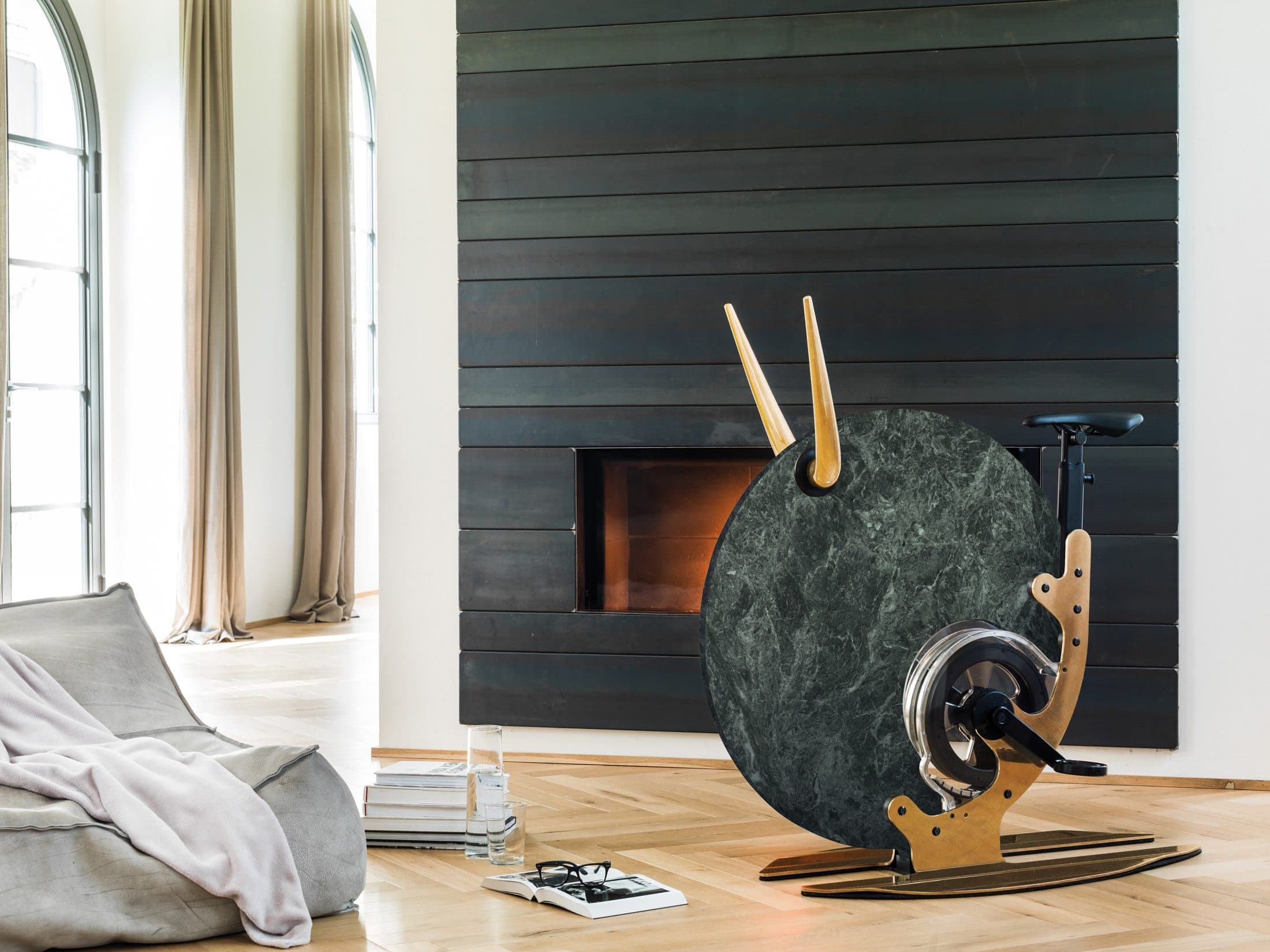 Bicicleta Ciclotte Marble con disco de mármol verde veteado y manubrios y estructura lateral en tono madera, junto a una chimenea encendida en un muro de madera oscura; a la izquierda, un puf con una manta y libros sobre el piso de duela.