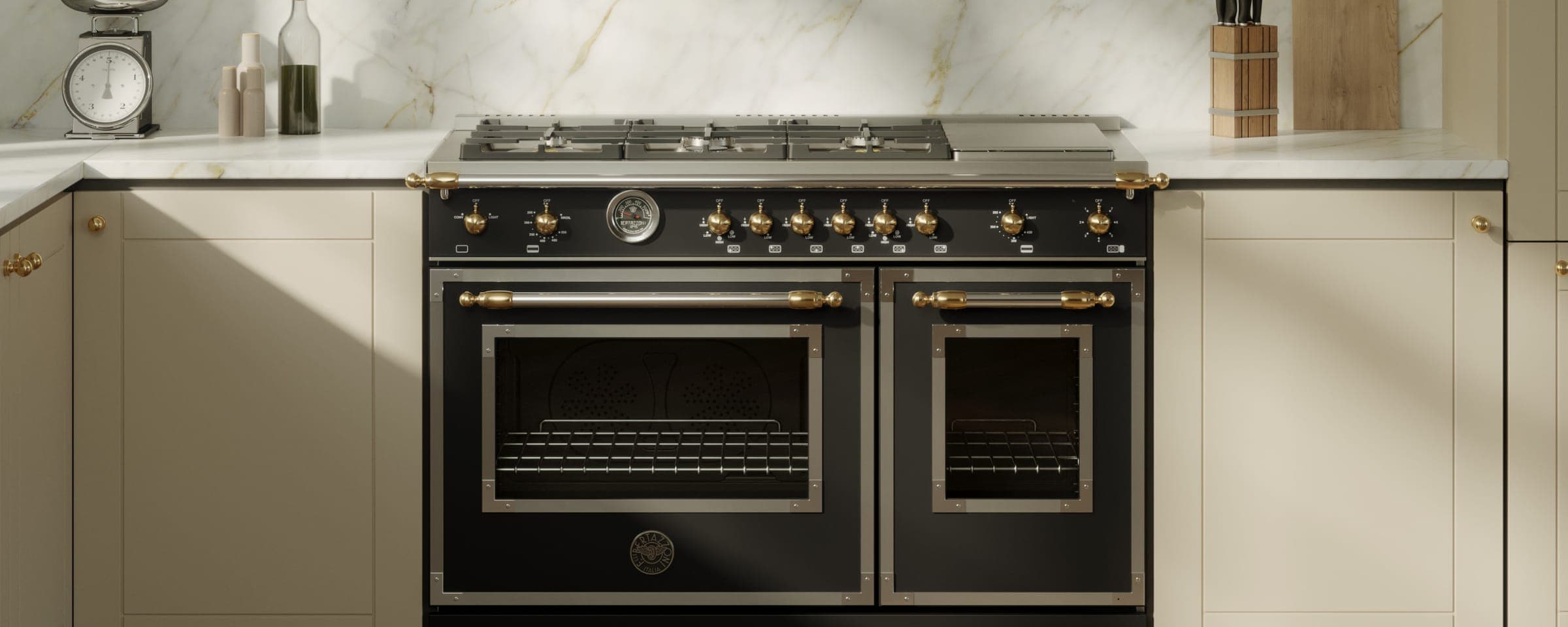 Estufa negra mate de dos hornos con quemadores y plancha, perillas doradas, termómetro y jaladeras cromadas con remates dorados, entre gabinetes color crema y un muro de mármol blanco con vetas doradas; a la izquierda, una báscula de cocina y botellas.