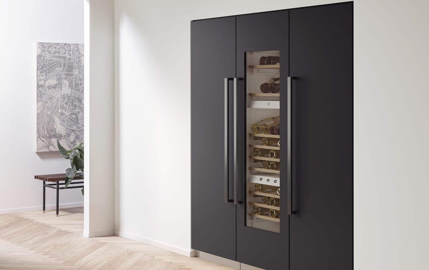 Tres columnas empotradas gris oscuro mate con jaladeras verticales largas; la del centro es una cava con puerta de vidrio y botellas sobre repisas de madera. Al lado, un pasillo blanco con piso de madera en espiga, una banca, una planta y un cuadro.