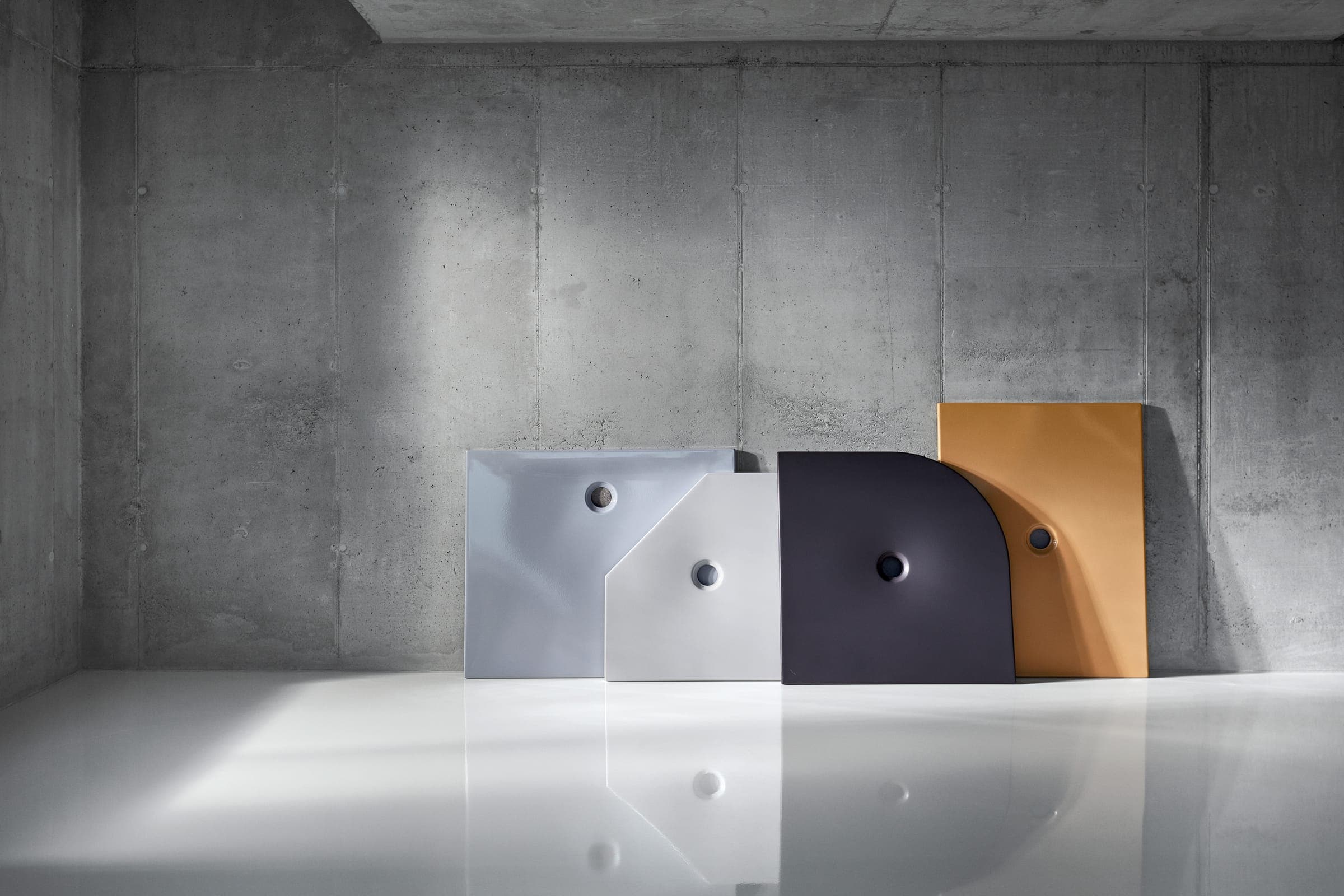 Cuatro platos de regadera, gris azulado, blanco, gris muy oscuro y ocre, de formas rectangular, pentagonal y de cuarto de círculo, recargados contra un muro de concreto aparente sobre un piso blanco brillante.