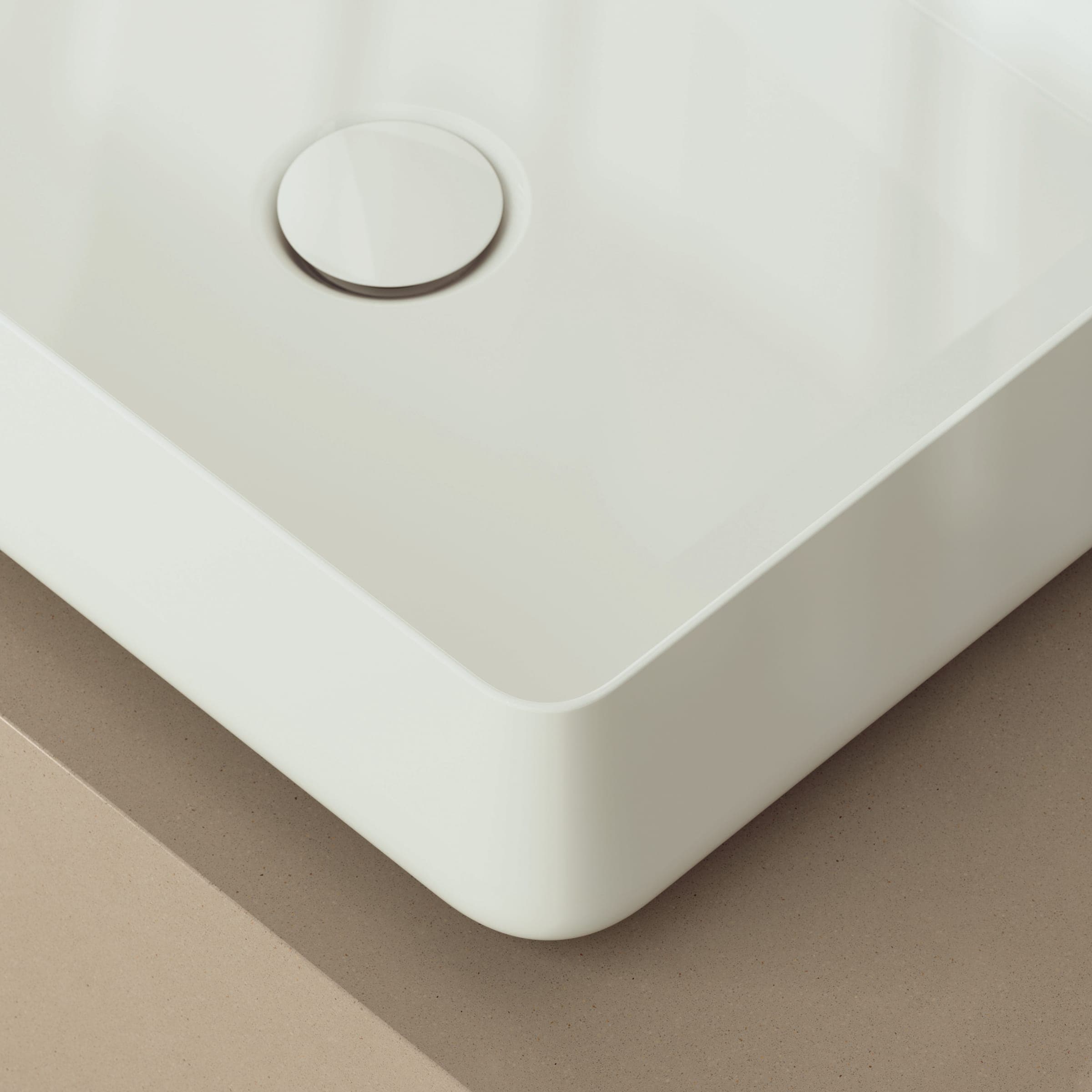 Acercamiento a la esquina de un lavabo rectangular blanco y brillante, de borde delgado, con su tapa de desagüe, sobre una cubierta beige.