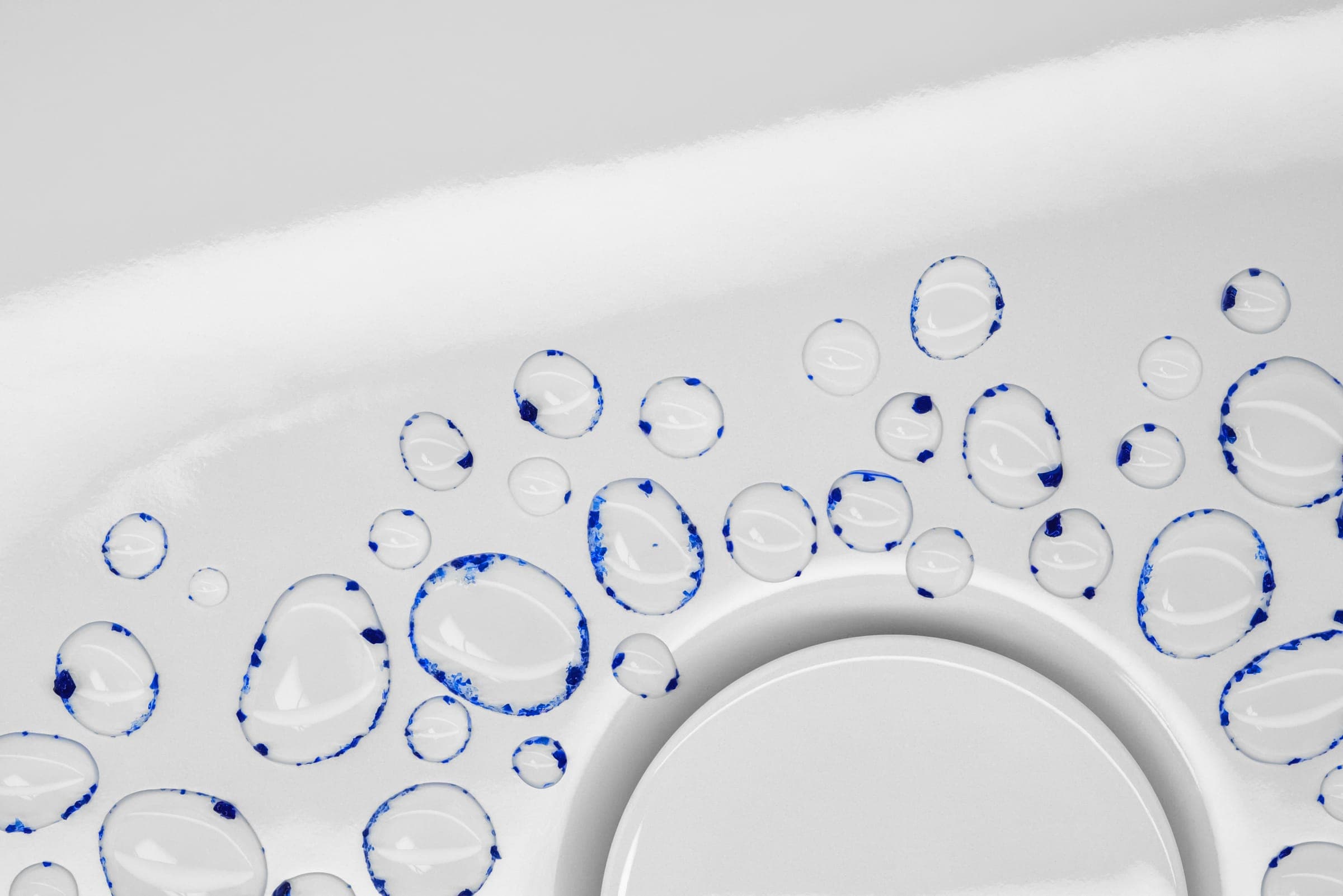 Gotas de agua con pigmento azul en los bordes sobre una superficie blanca, brillante y curva, junto a la tapa redonda de un desagüe.