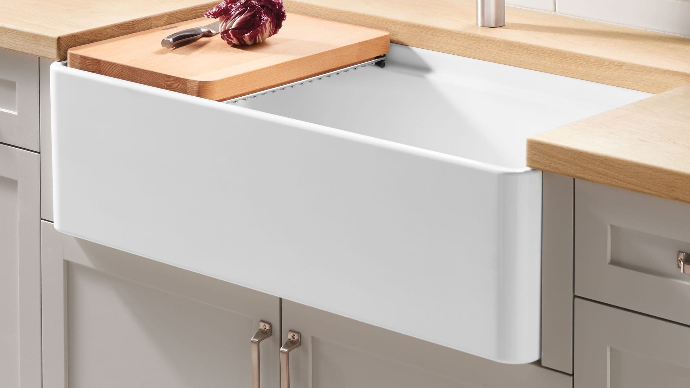 Tarja farmhouse blanca de frente expuesto entre cubiertas de madera clara y muebles grises con jaladeras metálicas; sobre la tina, una tabla de madera con un cuchillo y un radicchio.