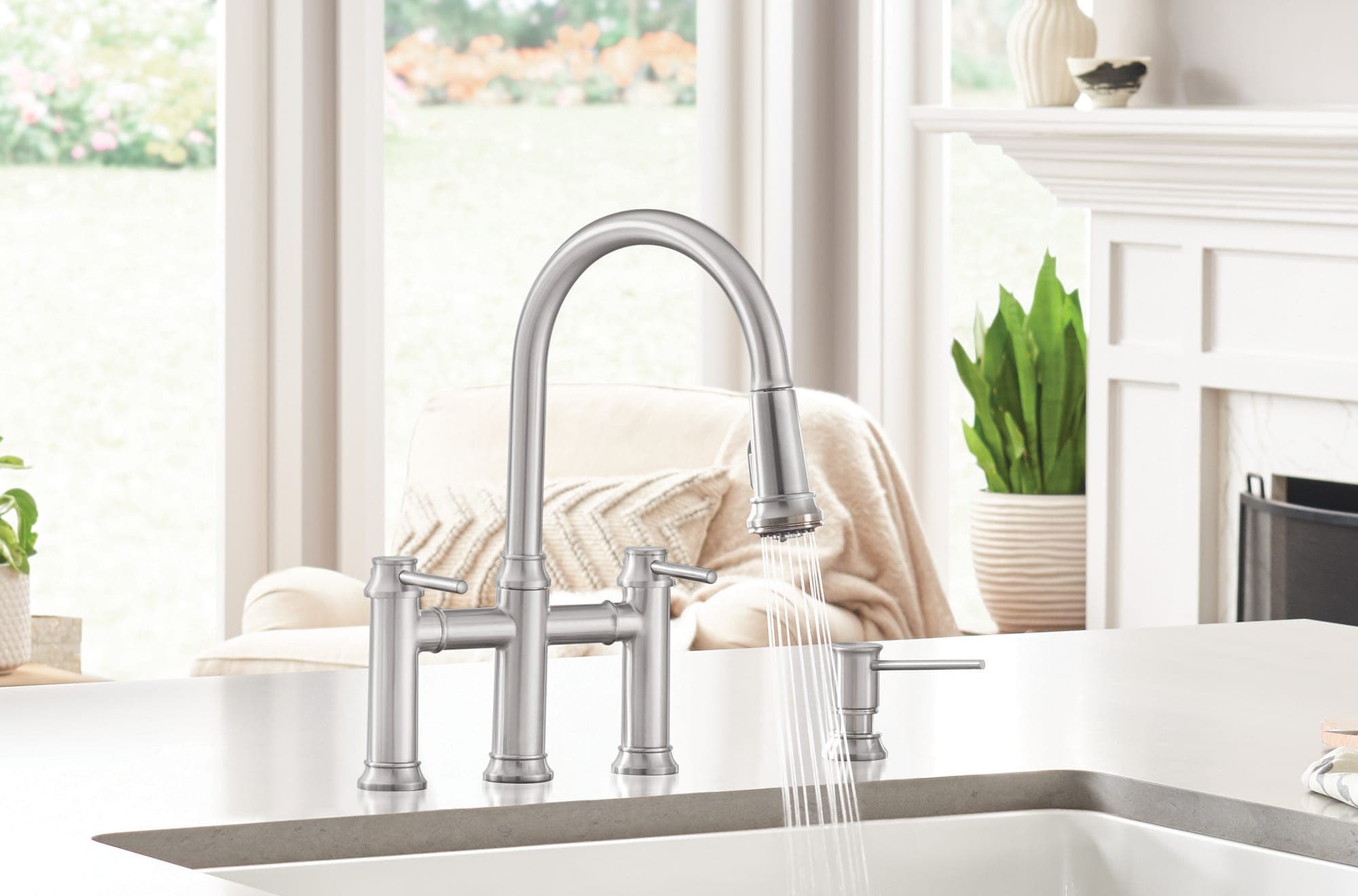 Mezcladora de puente de acero con regadera extraíble que deja caer agua en una tarja blanca, junto a un dispensador de jabón; al fondo, una sala luminosa con ventanales, un sillón claro y una chimenea blanca.