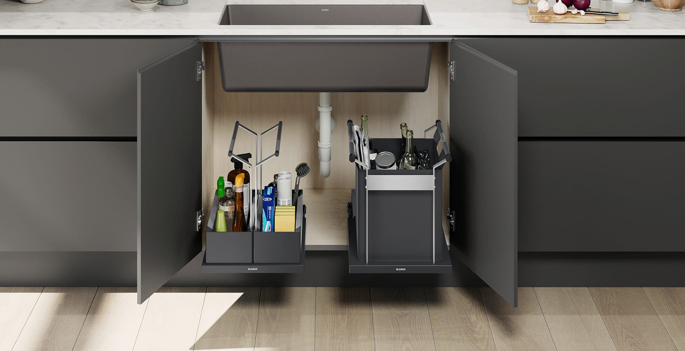 Mueble gris oscuro abierto bajo una tarja gris: a la izquierda, un organizador extraíble con productos de limpieza, esponjas y un cepillo; a la derecha, un bote extraíble con botellas de vidrio, una lata y periódico.