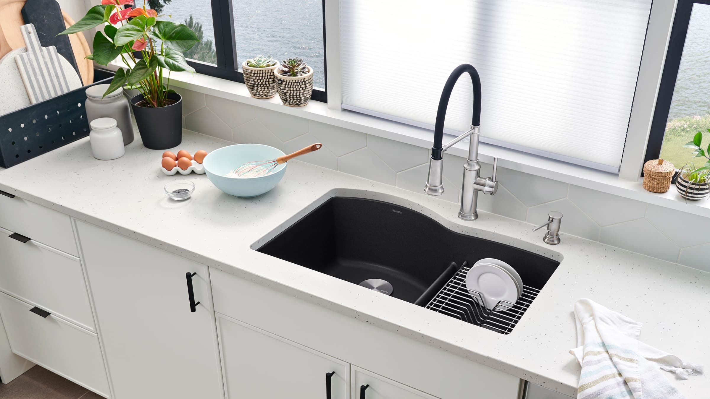 Cocina blanca junto a una ventana: tarja negra bajo cubierta con dos tinas desiguales y divisor bajo, una rejilla con platos, mezcladora de acero con manguera negra y dispensador de jabón; en la cubierta, huevos, un tazón con batidor y una planta.