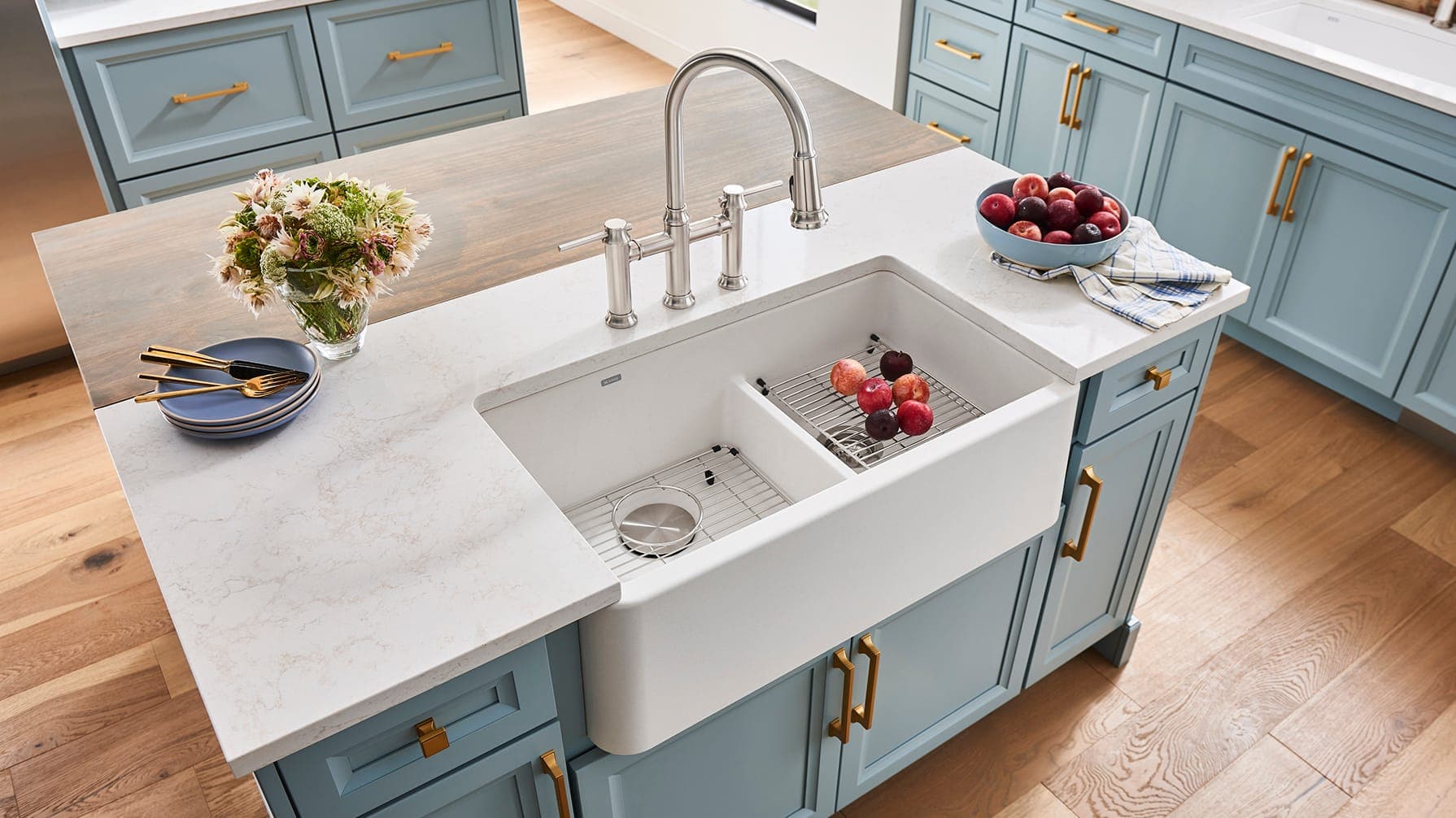 Isla de muebles azul claro con jaladeras doradas y cubierta blanca veteada: tarja farmhouse blanca de dos tinas con divisor bajo, rejillas de acero con fruta y una mezcladora de puente de acero; a los lados, un florero con flores, platos azules con cubiertos dorados y un tazón con fruta.