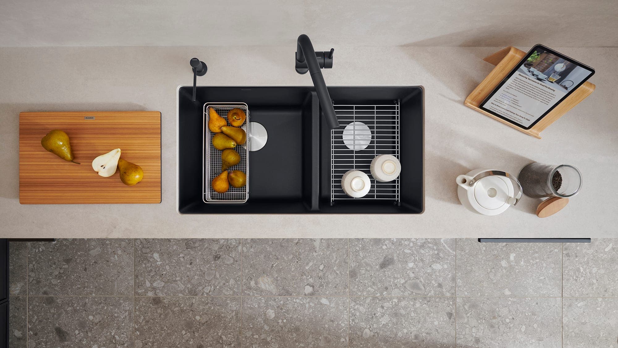 Vista cenital de una tarja negra de dos tinas desiguales: en una, un colador de malla con peras; en la otra, una rejilla con tazas blancas; en la cubierta clara, mezcladora y dispensador negros, una tabla de madera con peras, una tetera, un frasco y una tableta.
