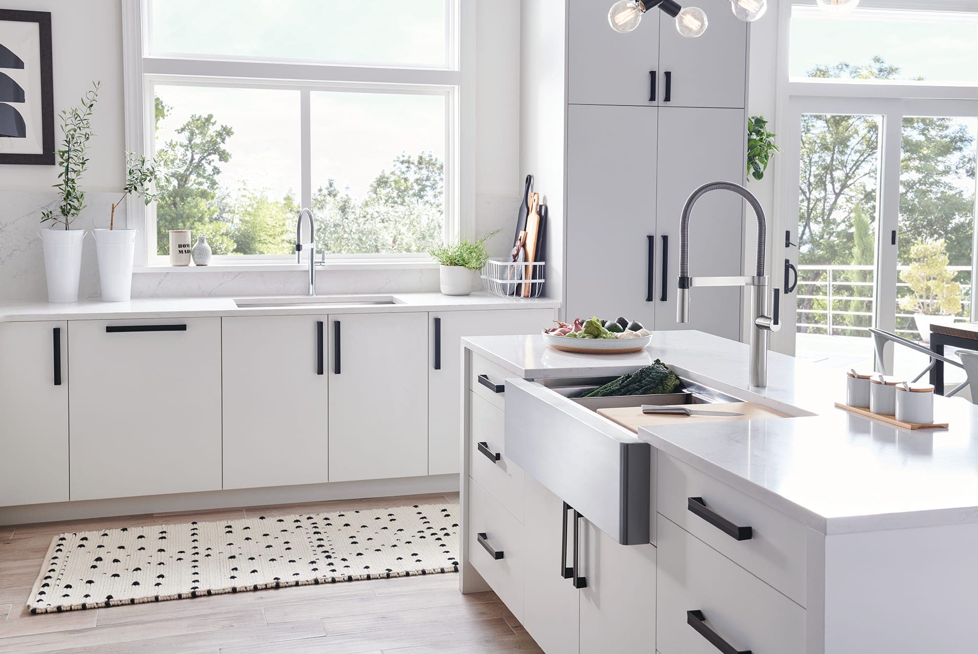 Cocina blanca con ventanales: en la isla, una tarja farmhouse de acero inoxidable de frente expuesto con una tabla de madera y col rizada en un colador, y una mezcladora semiprofesional de acero; al fondo, otra tarja bajo la ventana.