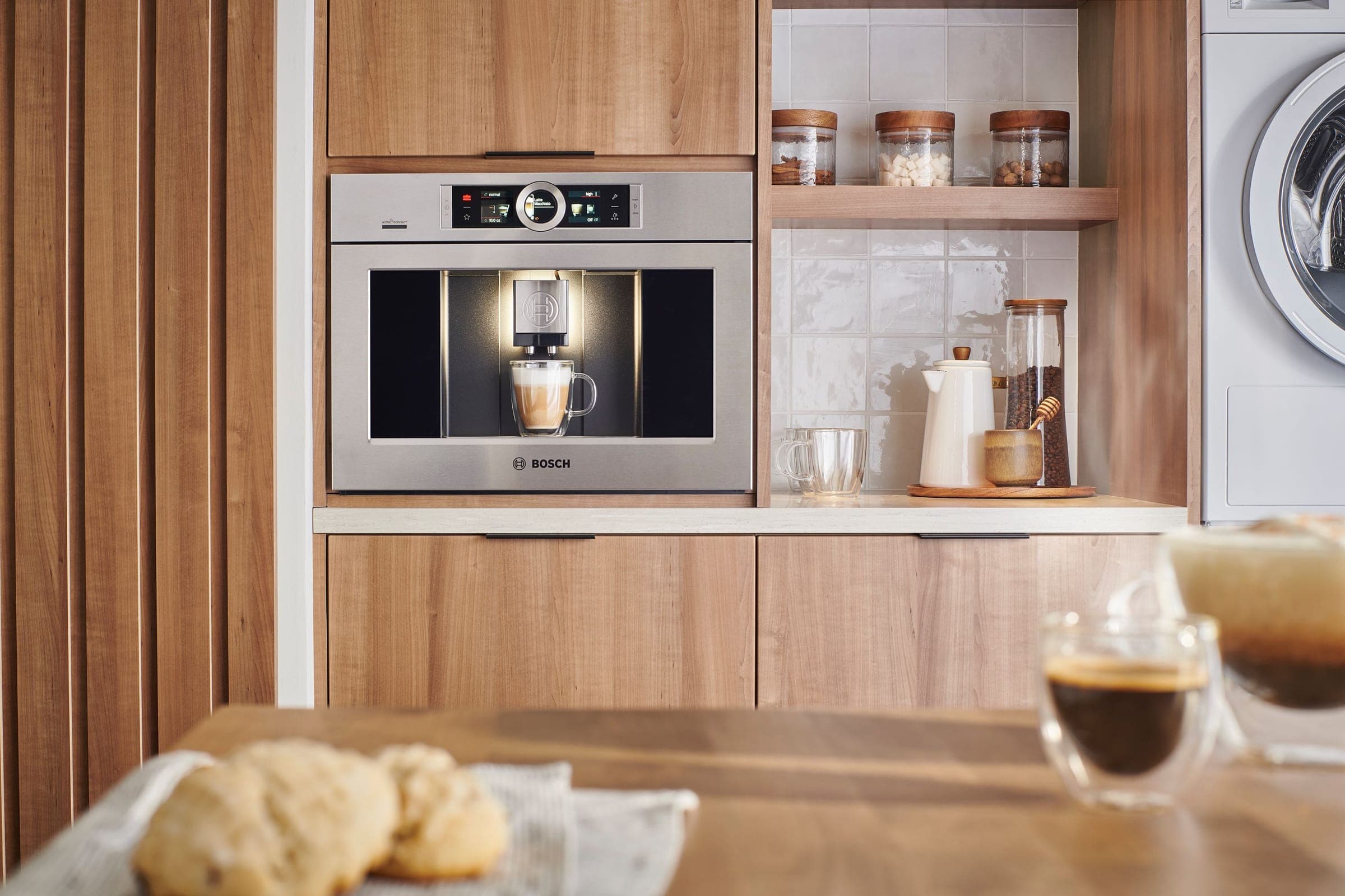 Cafetera empotrable de acero inoxidable con logotipo BOSCH sirviendo un café con leche en un vaso de vidrio, en un mueble de madera con repisa de frascos y azulejo blanco; en primer plano, una mesa de madera con panecillos y vasos de vidrio con café.