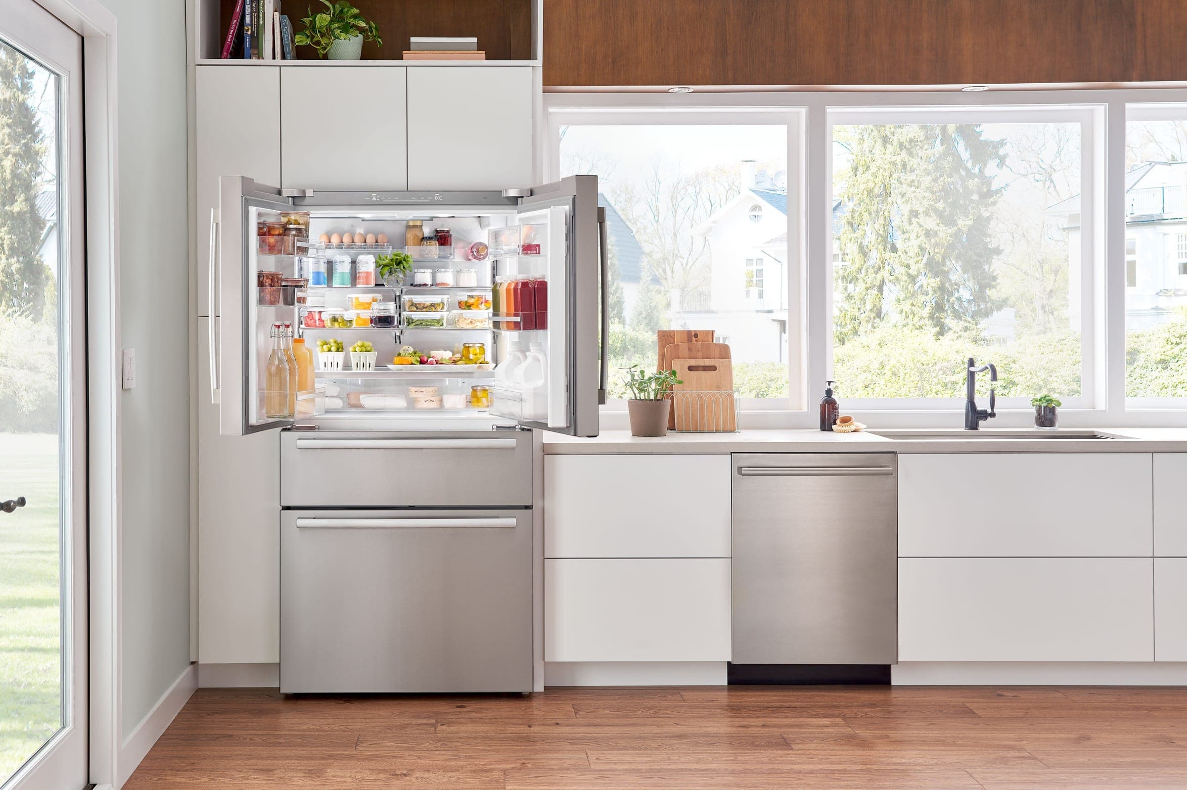 Refrigerador de acero inoxidable de puertas francesas con dos cajones congeladores, abierto y lleno de alimentos, junto a un lavavajillas de acero inoxidable, en una cocina de gabinetes blancos con ventanales hacia un jardín, llave negra y piso de madera.