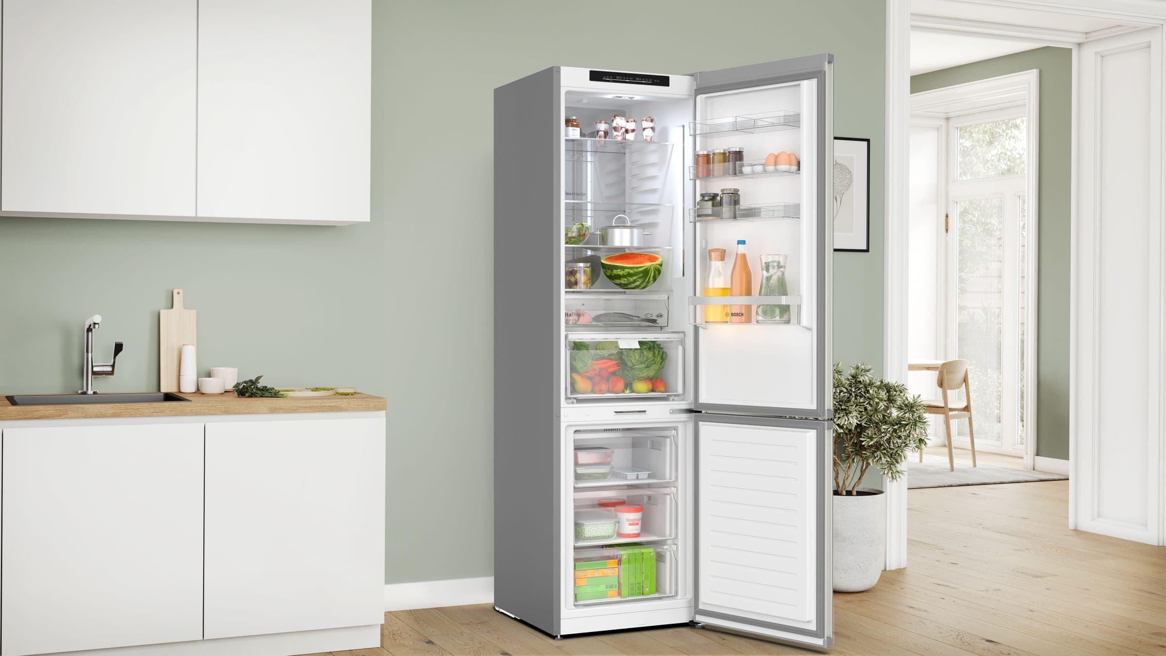 Refrigerador angosto de acero inoxidable con congelador inferior, con ambas puertas abiertas y lleno de alimentos, en una cocina de muros verde salvia con gabinetes blancos, cubierta de madera y piso de duela.