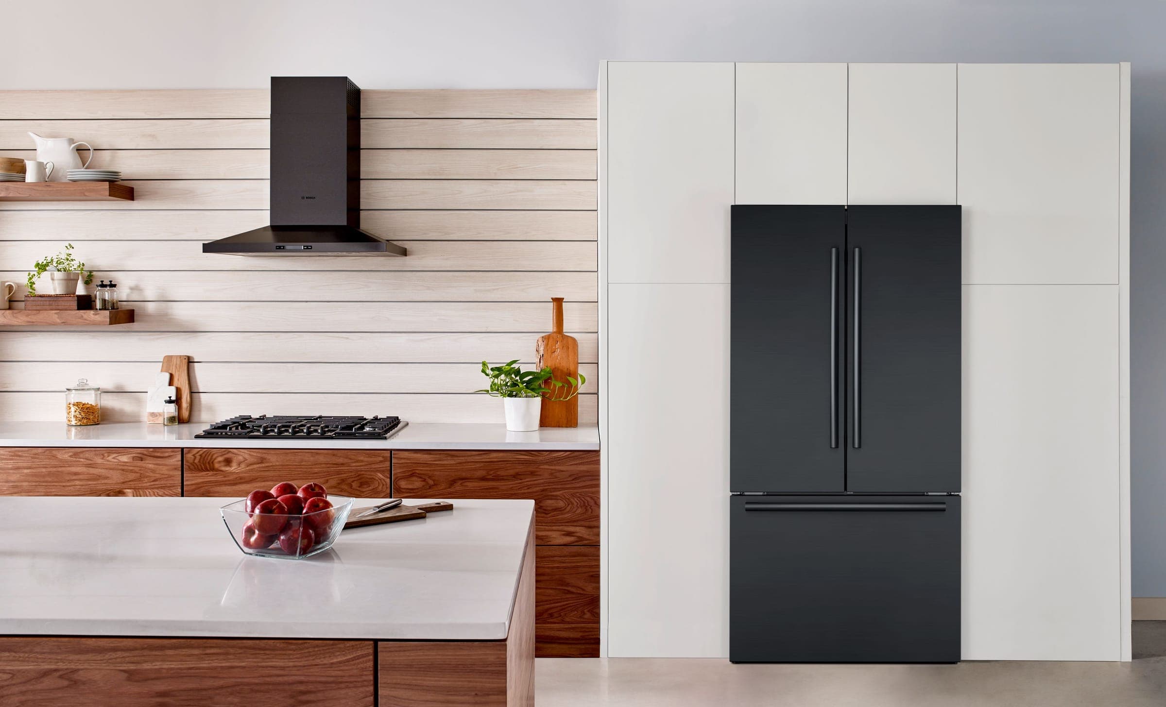 Refrigerador de puertas francesas en acero inoxidable negro, cerrado, integrado en un muro de gabinetes blancos; a la izquierda, una campana de chimenea negra sobre una cubierta de gas, un muro de listones de madera clara con repisas y una isla con un tazón de manzanas.