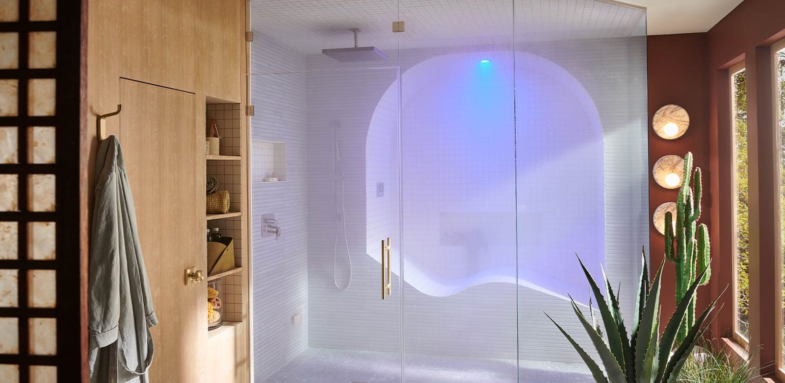 Regadera de vapor con cancel de vidrio, mosaico blanco y una banca curva bajo un arco, llena de vapor iluminado en azul y violeta desde el techo; regadera cuadrada de techo, muebles de madera, bata colgada, agave y cactus.