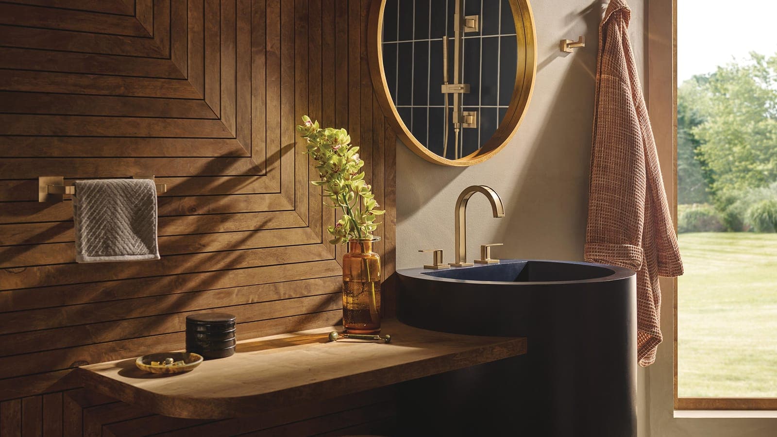Lavabo cilíndrico negro con llave dorada de caño curvo y dos manijas junto a un muro de madera con líneas en ángulo; espejo redondo dorado que refleja una regadera de mano en barra, orquídeas en un florero ámbar, bata colgada y ventana al jardín.