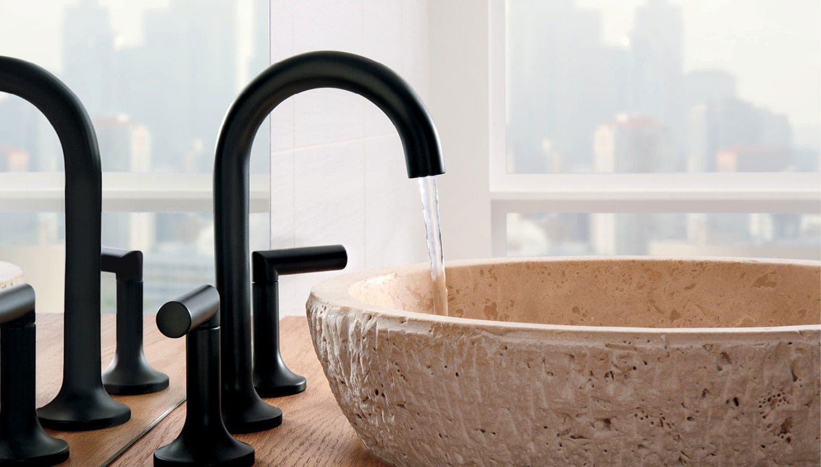 Llave de lavabo negra mate de cuello alto y dos manijas vertiendo agua en un lavabo de piedra de textura rugosa; a la izquierda, su reflejo en un espejo y, al fondo, una ventana con edificios difuminados.