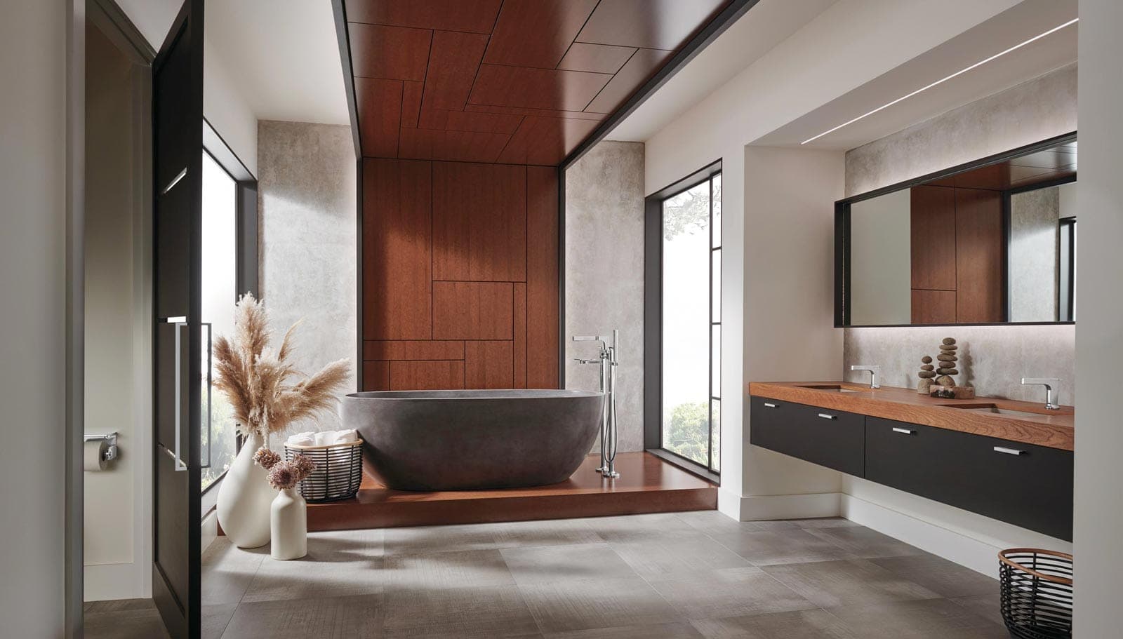 Baño con tina exenta gris oscuro sobre una tarima de madera y llave de piso cromada; muro y plafón de paneles de madera rojiza, ventanales y, a la derecha, mueble flotante con dos lavabos y un espejo largo.