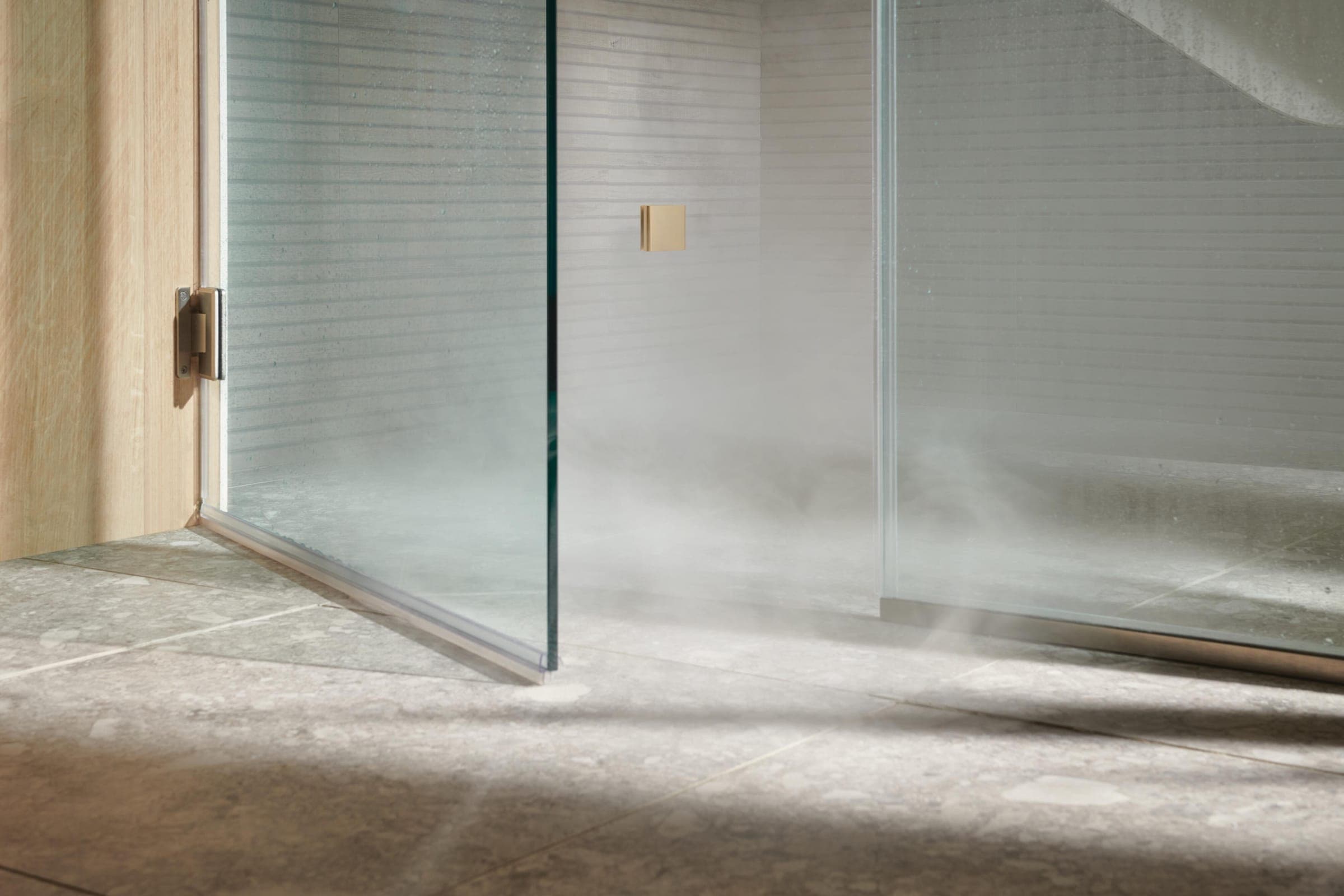 Parte baja de una regadera de vapor: puerta de vidrio abierta sobre su bisagra, vapor a ras de un piso de piedra gris y un cabezal de vapor cuadrado dorado en el muro de mosaico blanco.