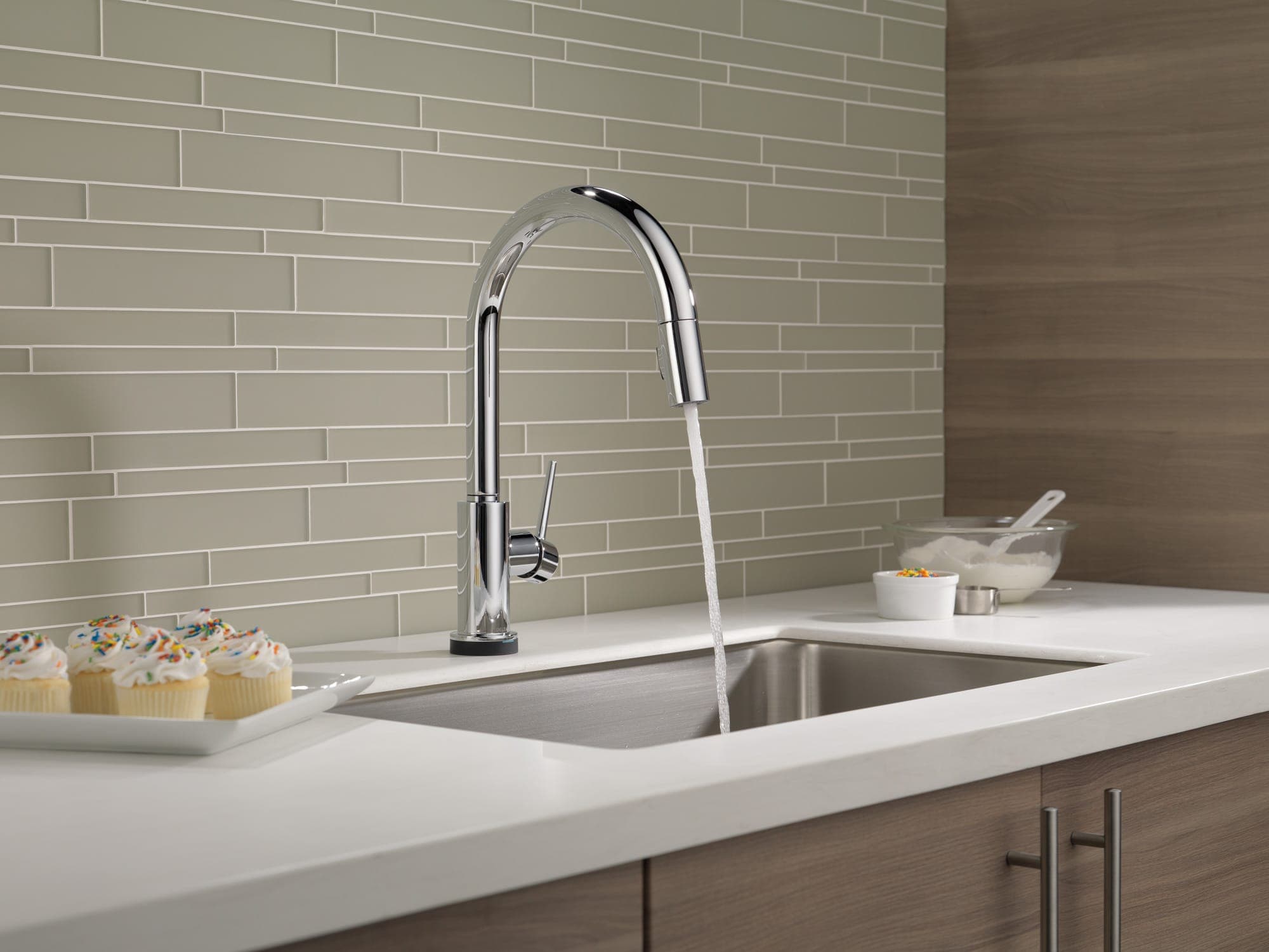 Monomando extraíble cromado de cuello alto con chorro de agua sobre una tarja de acero inoxidable bajo cubierta; cubierta blanca, muro de loseta lineal beige, gabinetes de madera y cupcakes al lado.
