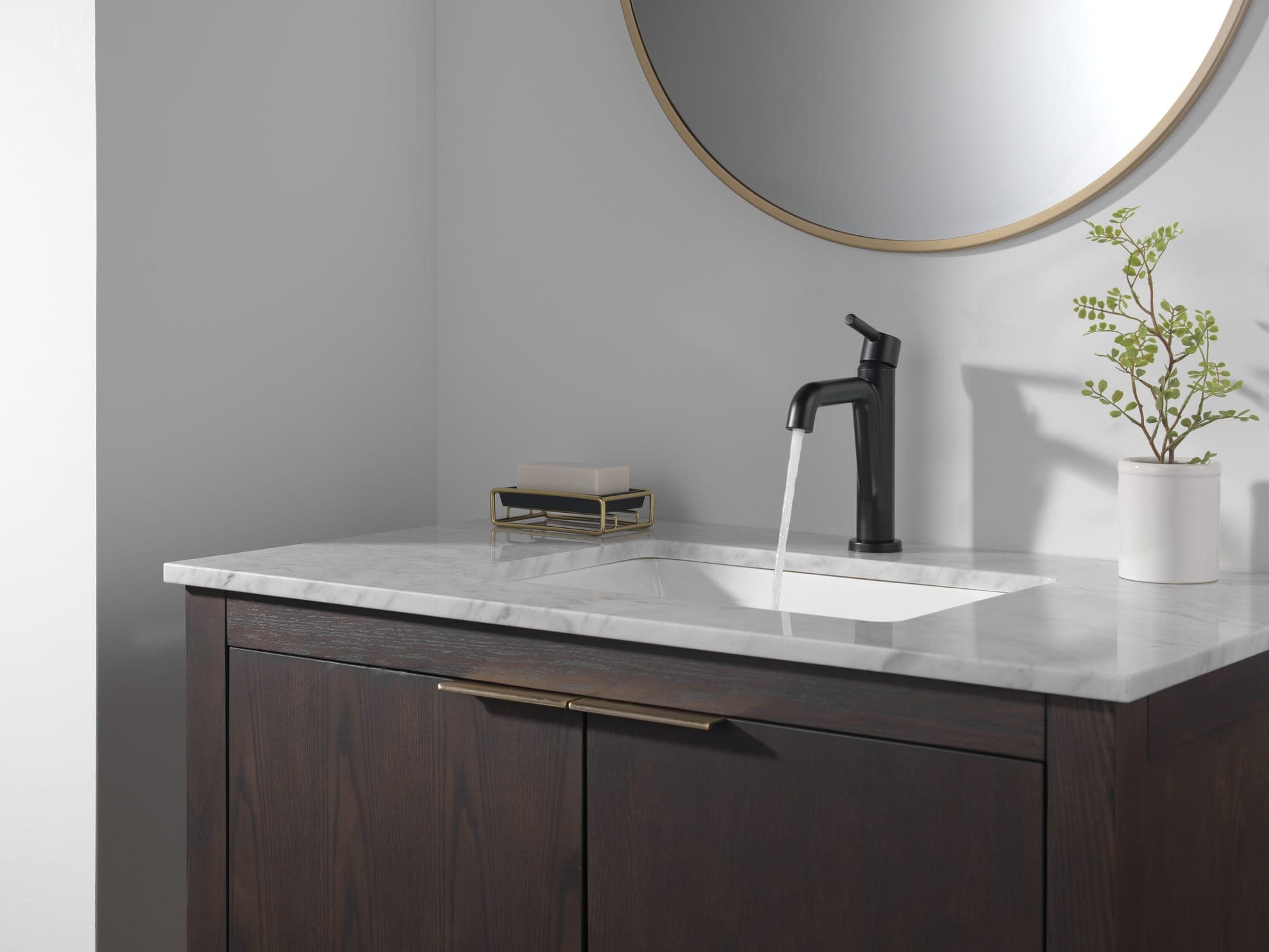 Monomando negro mate con agua corriendo sobre un lavabo bajo cubierta de mármol blanco; mueble de madera oscura, espejo redondo de marco dorado, jabonera y una planta.