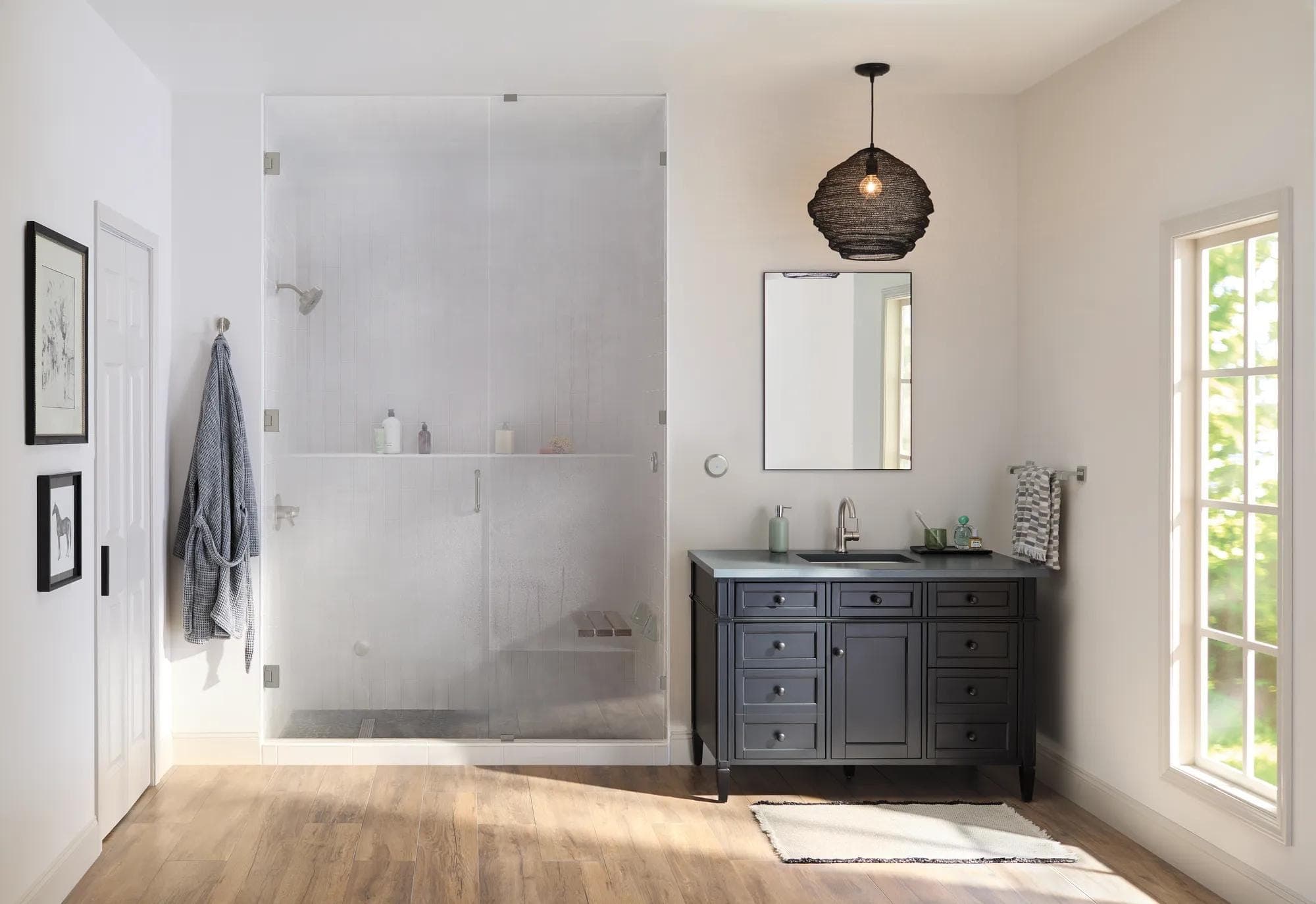 Baño luminoso con una regadera cerrada por cancel de vidrio empañado por el vapor y un asiento de tablas de madera adentro; a la derecha, tocador oscuro con llave plateada, espejo y lámpara colgante; piso de madera.