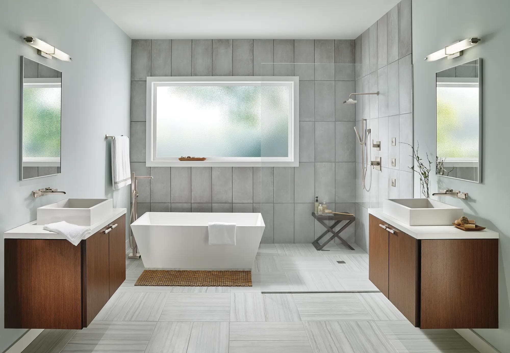 Baño simétrico con una tina exenta rectangular al centro y su llenador de piso a un lado, dos muebles flotantes de madera con lavabos de sobreponer y llaves de muro, y regadera abierta con rociadores corporales; muros de loseta gris.