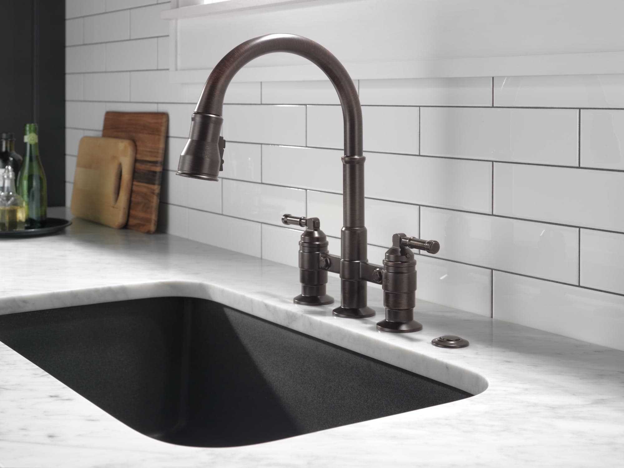 Llave de cocina tipo puente en bronce oscuro, con regadera extraíble y dos manerales de palanca, sobre cubierta de mármol blanco con tarja negra bajo cubierta; azulejo blanco tipo metro y tablas de picar.