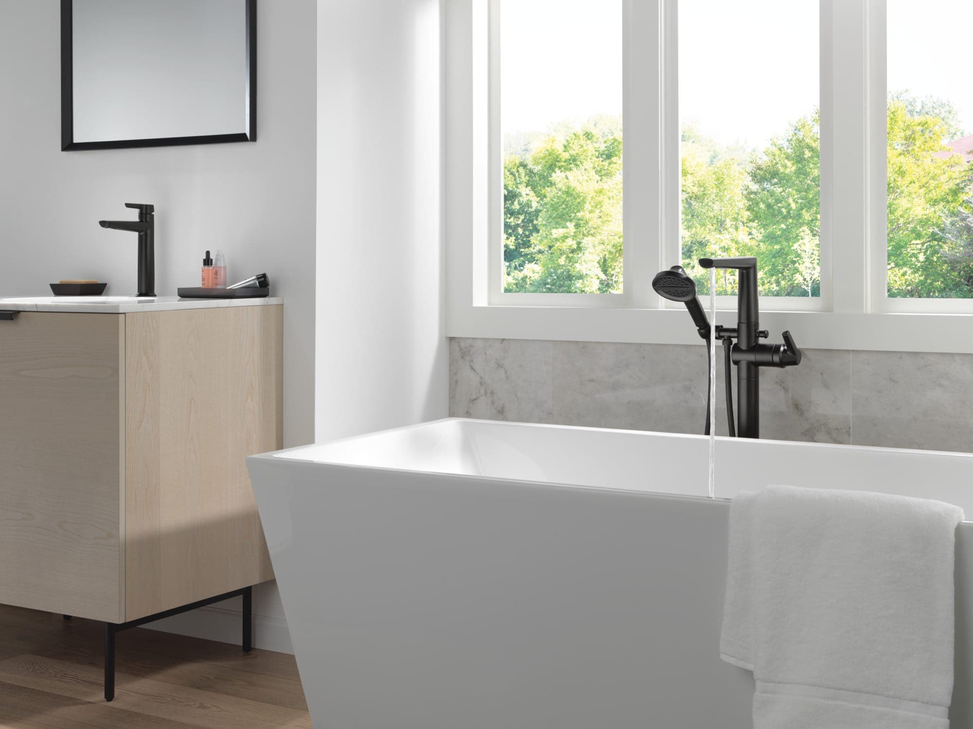 Llenador de tina de piso negro mate con regadera de mano vertiendo agua en una tina exenta rectangular blanca; a la izquierda, mueble de madera clara con monomando negro; ventanal con árboles.