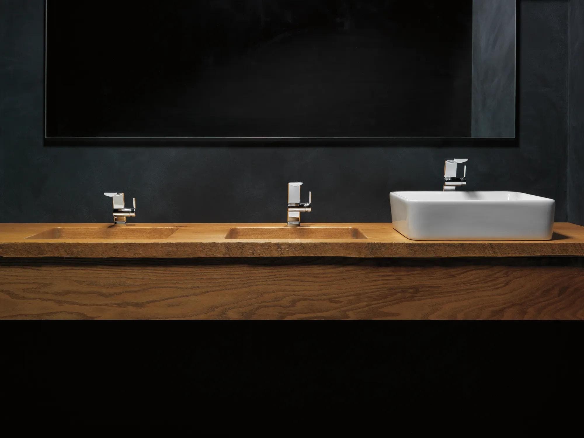 Tres monomandos cromados de distinta altura sobre una cubierta de madera: dos con lavabos tallados en la madera y uno sobre un lavabo de sobreponer blanco; muro negro y espejo horizontal.