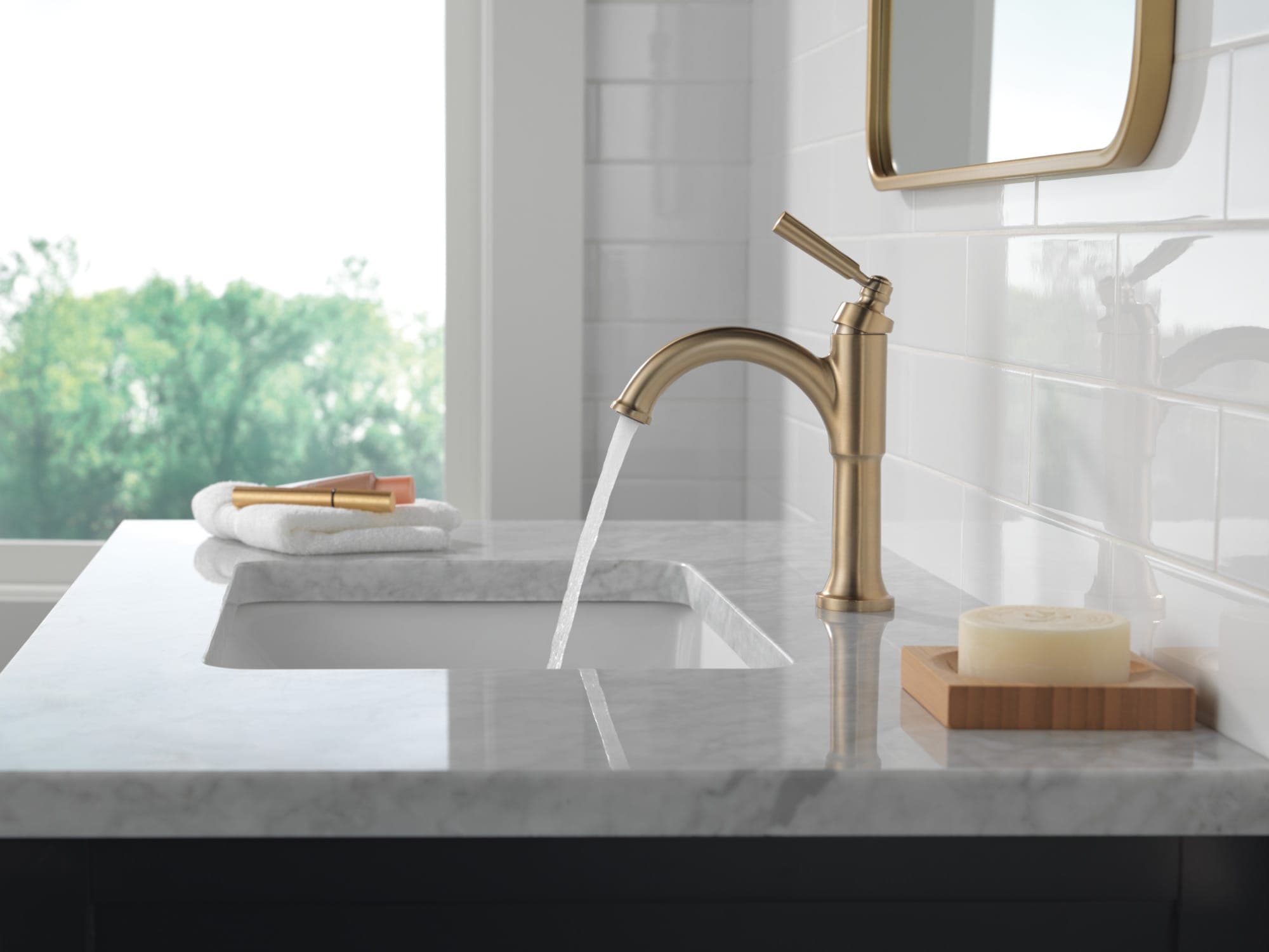 Monomando en tono dorado claro con palanca alta y agua corriendo sobre un lavabo bajo cubierta de mármol; azulejo blanco brillante, ventana con árboles, espejo de marco dorado y jabón sobre una base de madera.