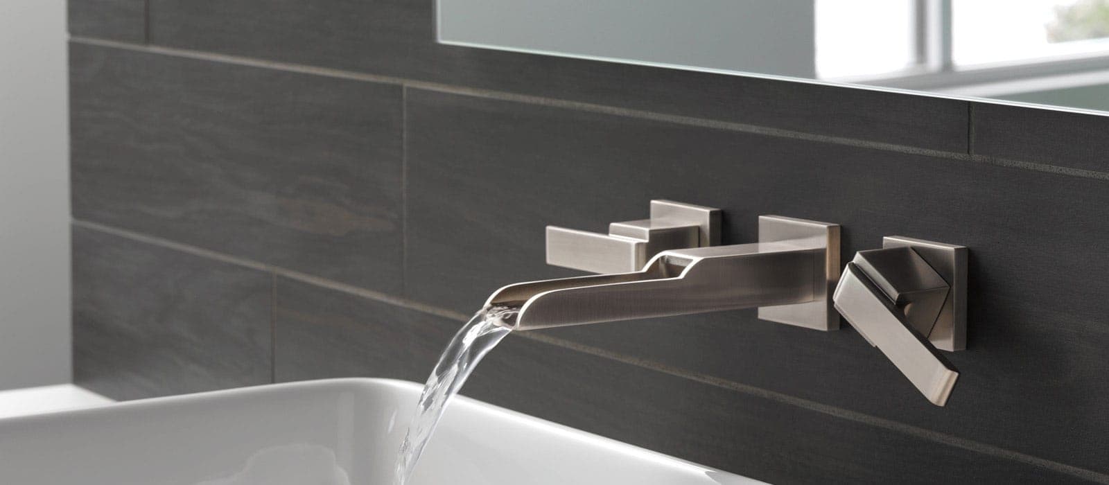 Llave de muro en acabado plateado satinado con caño abierto que vierte una lámina de agua sobre un lavabo blanco, con manerales de palanca a ambos lados; muro de loseta gris oscuro y espejo arriba.