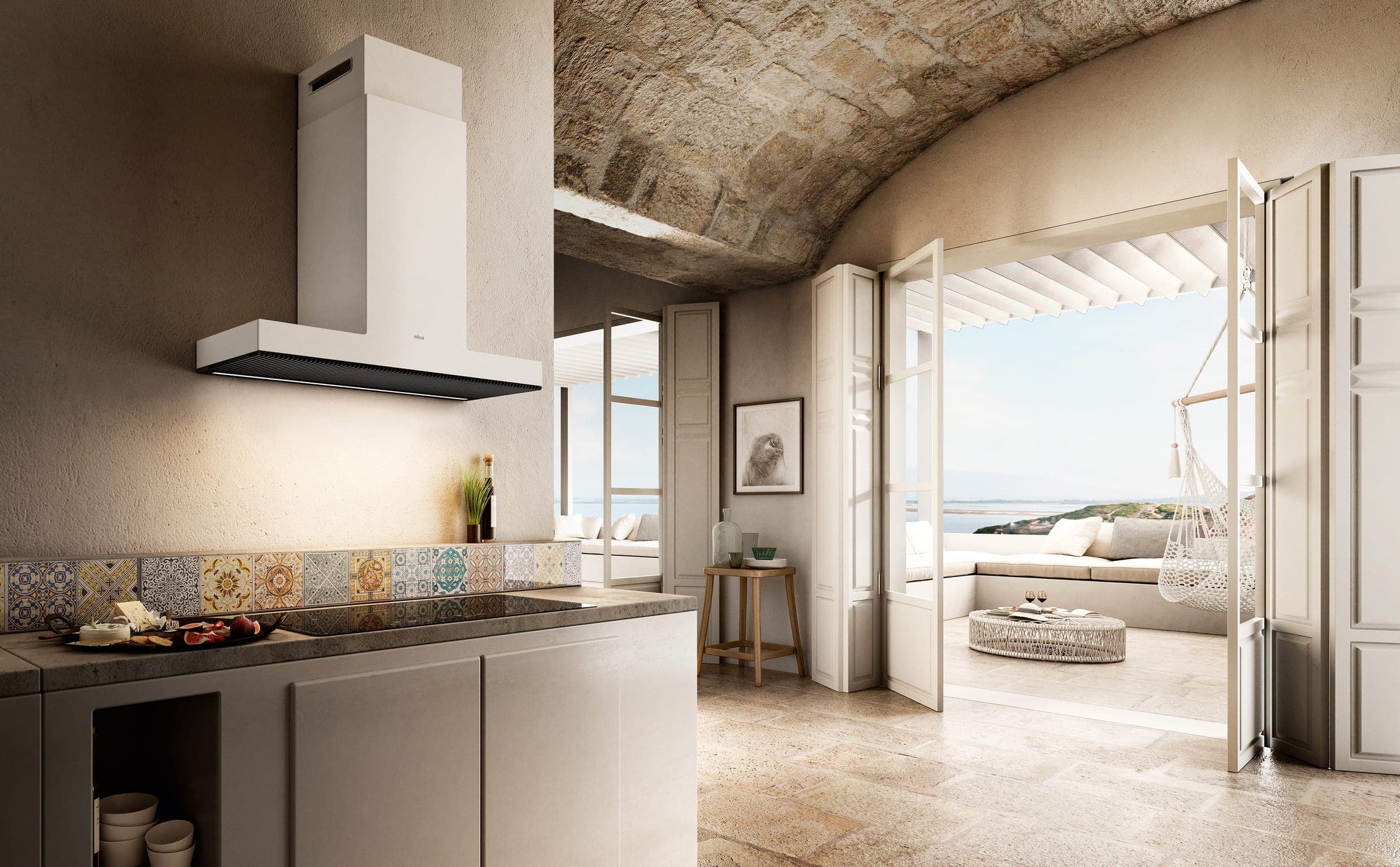 Campana de pared blanca con chimenea sobre una cubierta de cocción y un muro de azulejos de colores, en una cocina con techo abovedado de piedra; puertas abiertas a una terraza con vista al mar.
