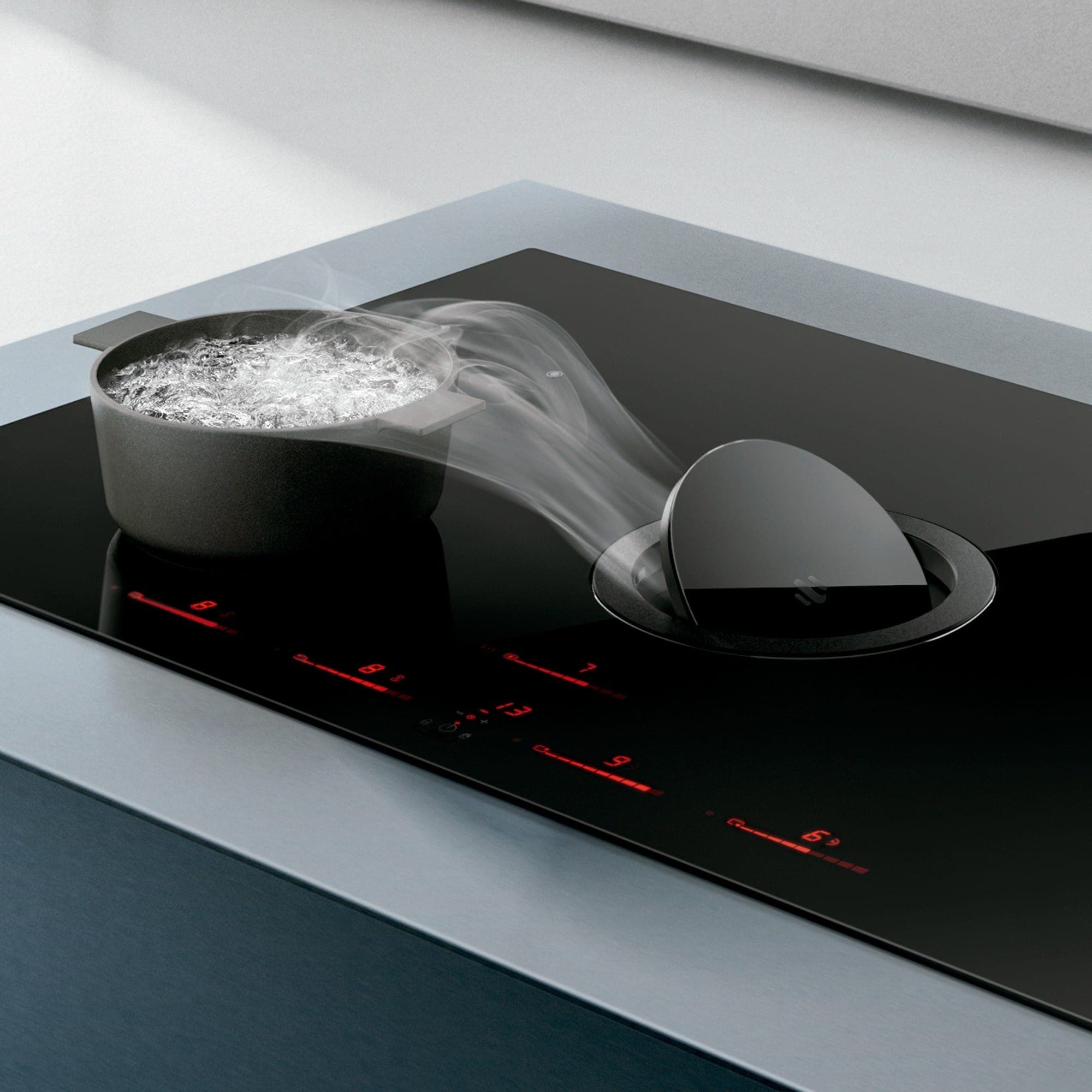 Acercamiento a una cubierta de inducción negra con borde de acero: el vapor de una olla con agua hirviendo se desvía hacia el extractor integrado, de compuerta circular levantada, junto a los indicadores rojos del control táctil.