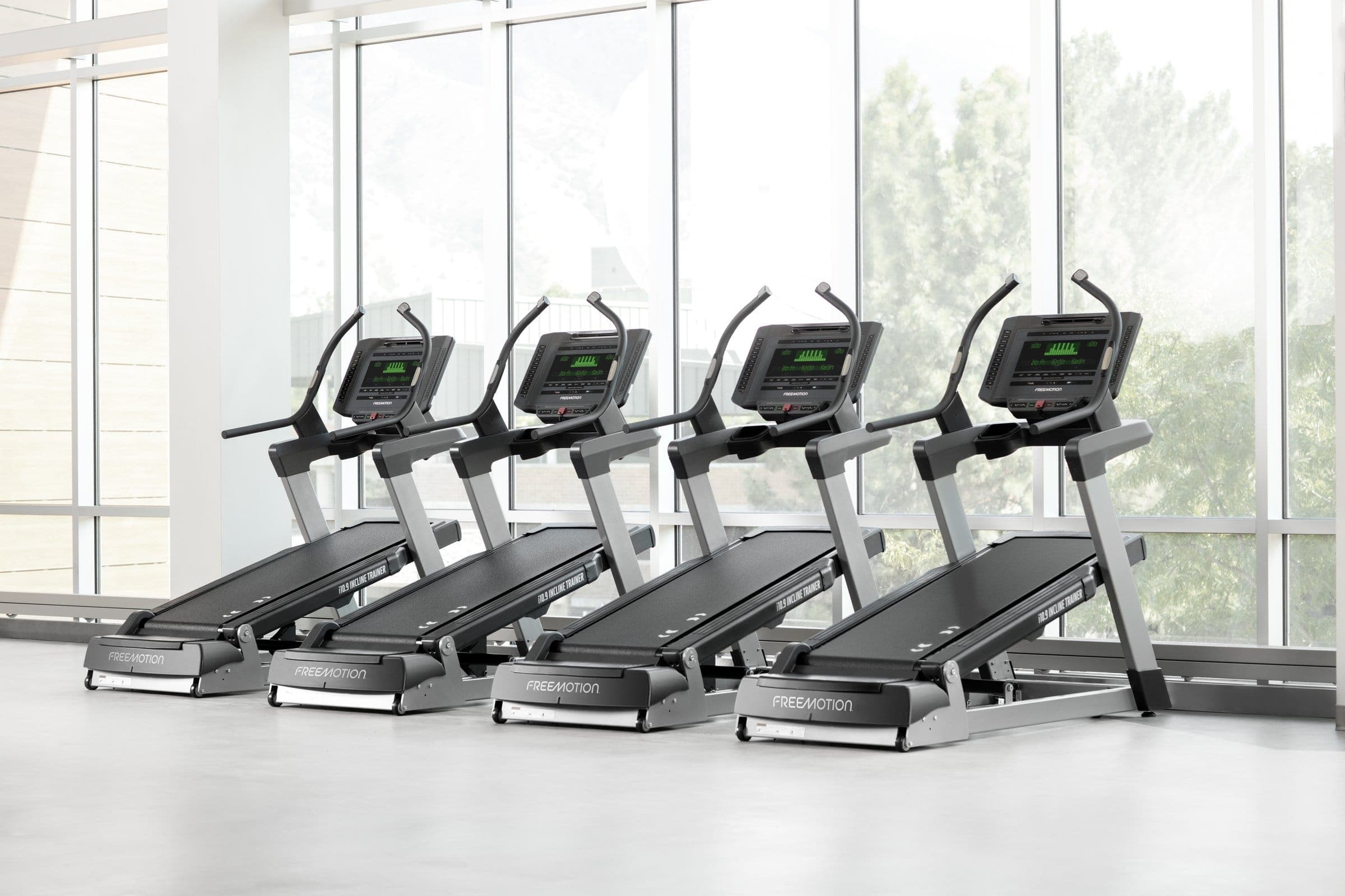 Cuatro Incline Trainers i10.9 de Freemotion alineados en diagonal frente a un ventanal con árboles, en una sala blanca y vacía.