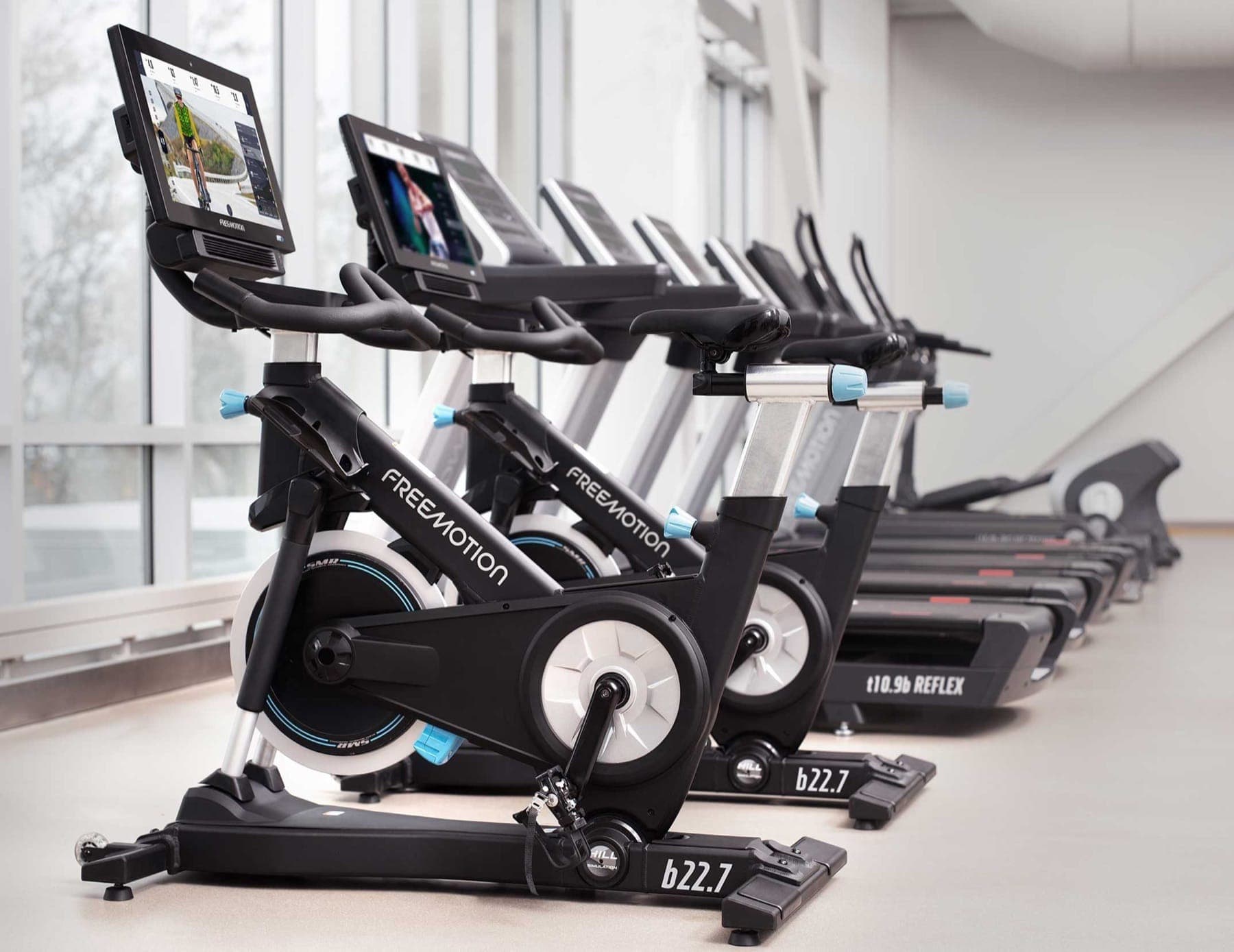 Fila de bicicletas CoachBike b22.7 de Freemotion con volante plateado, perillas azul claro y pantallas encendidas; al fondo, caminadoras t10.9b REFLEX junto a ventanales.
