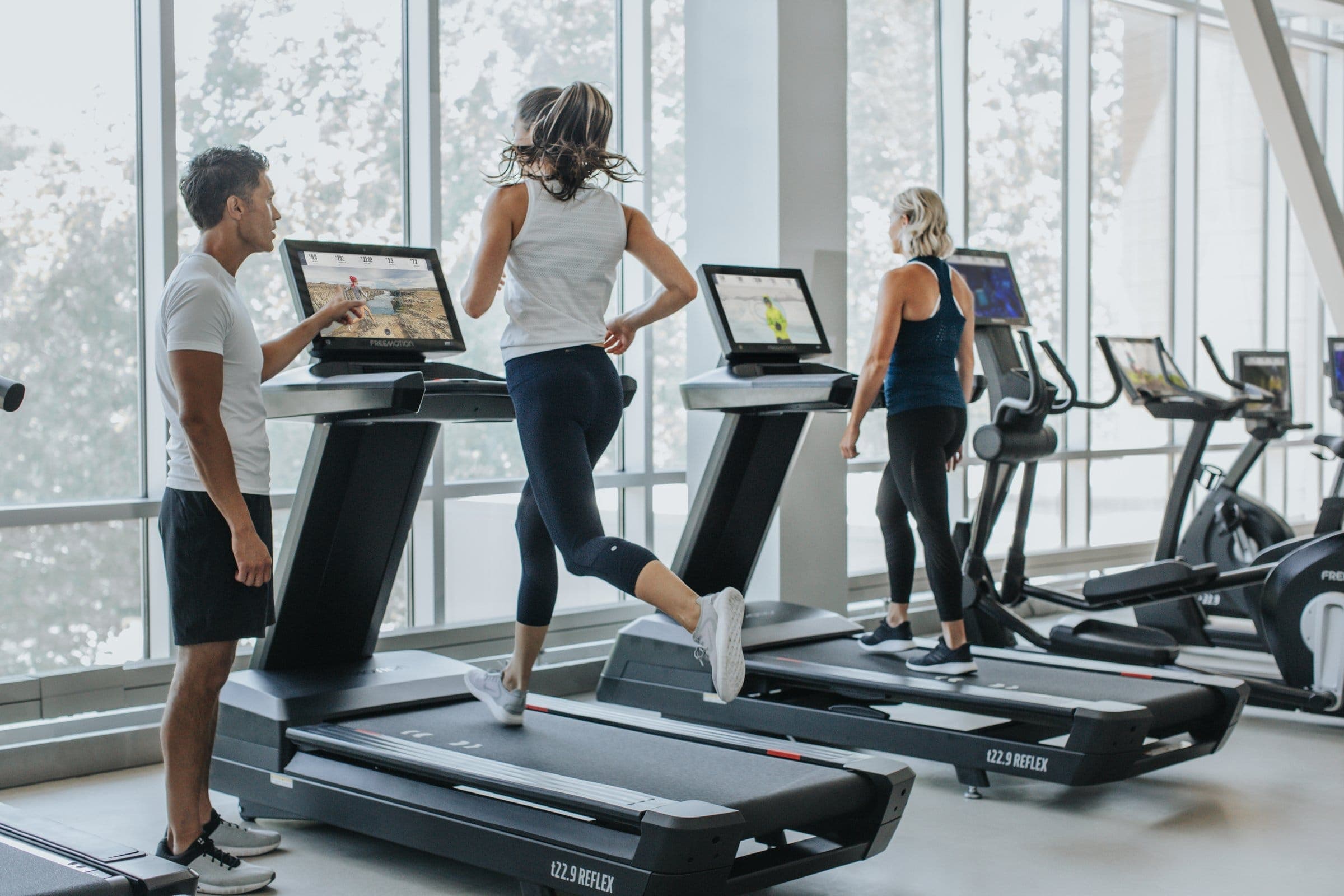 Caminadoras t22.9 REFLEX de Freemotion junto a un ventanal: un hombre señala la pantalla a una mujer que corre y otra mujer camina en la siguiente; al fondo, otras máquinas de cardio.