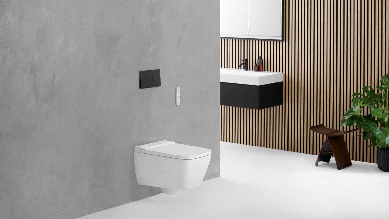 Inodoro con ducha AquaClean Sela, de líneas rectas, suspendido en un muro gris tipo concreto con placa actuadora negra y control remoto blanco; al fondo, muro de listones de madera, lavabo con mueble negro, un banco de madera y una planta.