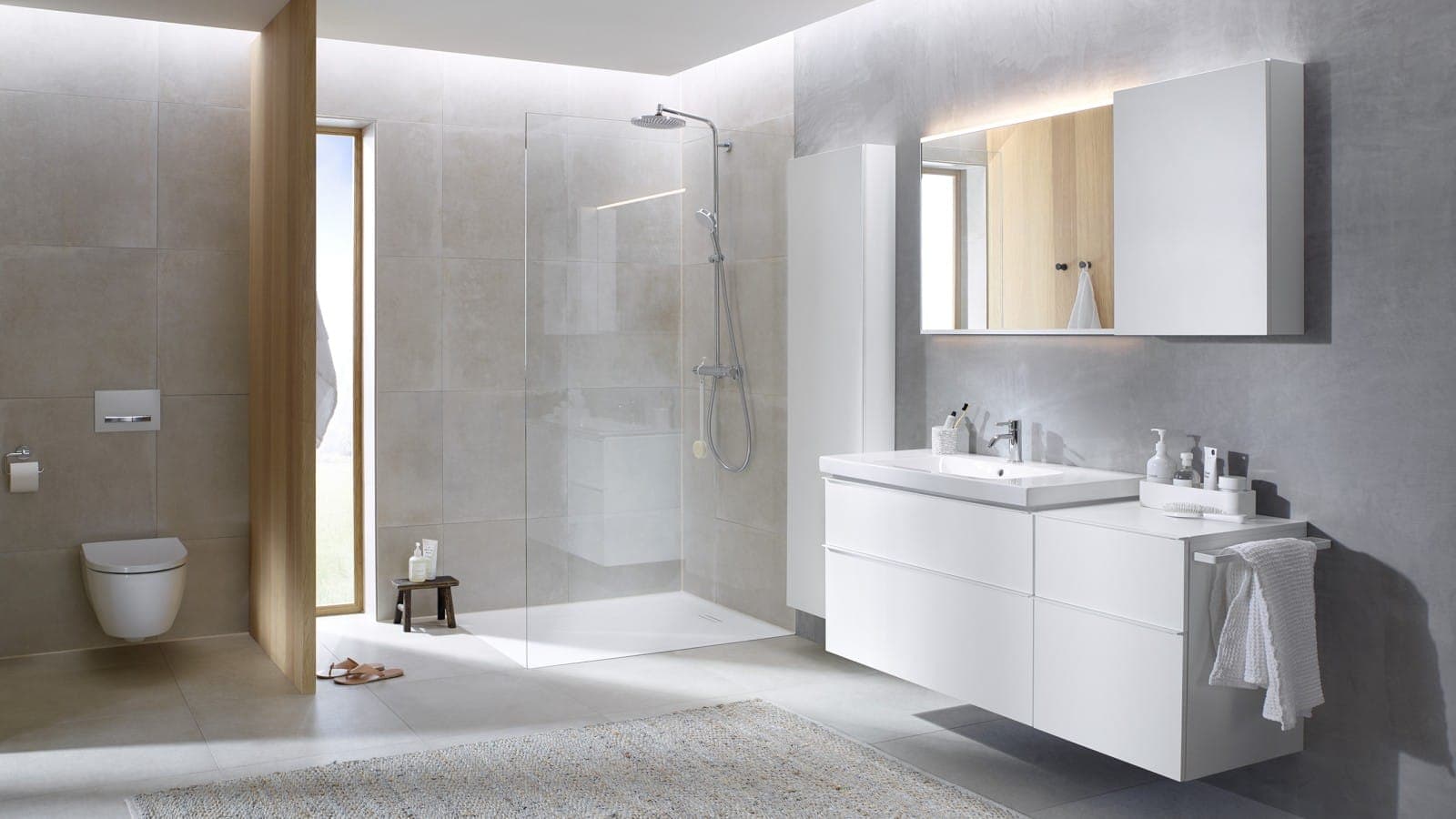 Baño en tonos gris claro con mueble de lavabo blanco de cajones, columna alta blanca y gabinete con espejo iluminado; a la izquierda, un inodoro suspendido y una regadera con cancel de vidrio.
