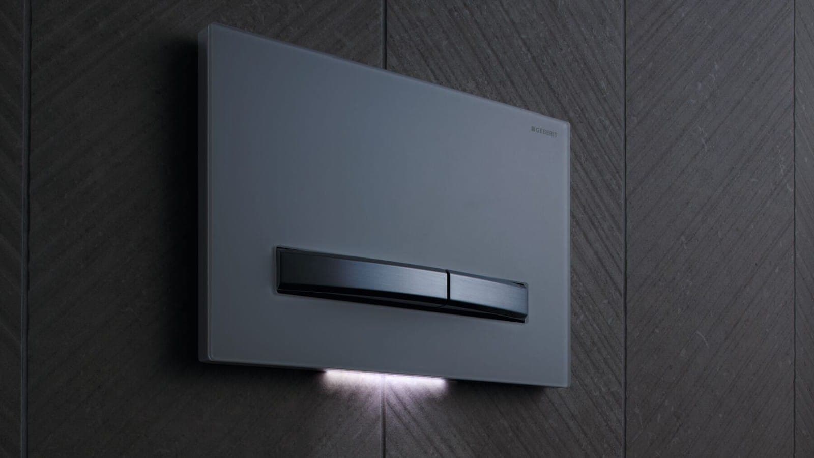Placa actuadora Sigma50 gris con dos botones oscuros de acabado brillante sobre un muro oscuro con textura diagonal; por debajo de la placa sale una luz blanca.