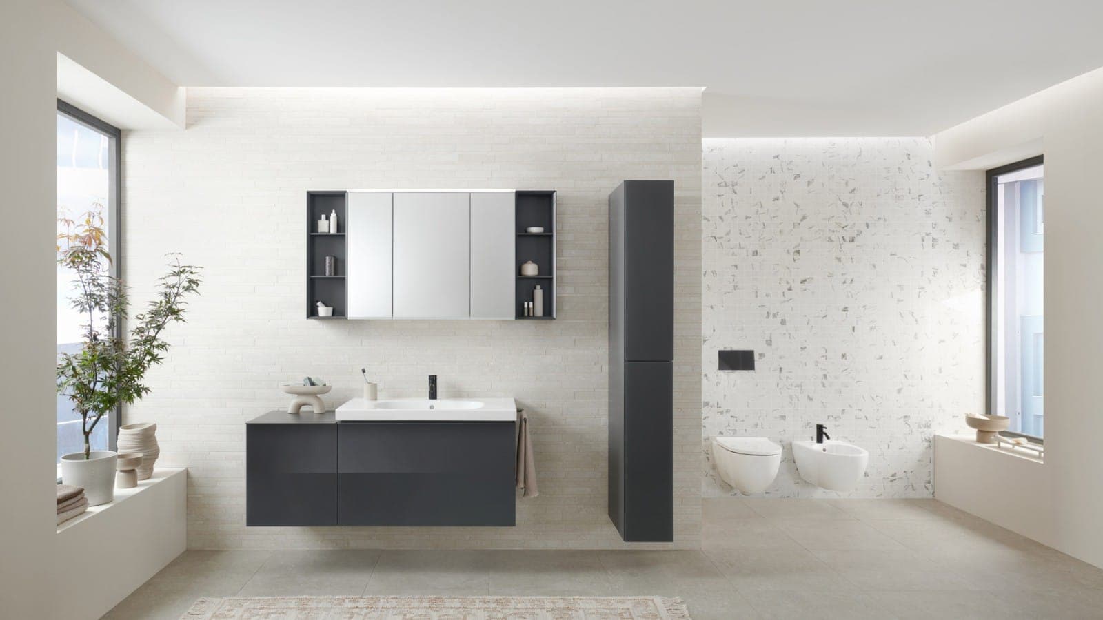 Baño claro con mueble de lavabo gris oscuro, gabinete con espejo con repisas abiertas y columna alta gris; al fondo, inodoro y bidé suspendidos sobre un muro de mosaico blanco y gris con placa negra; a los lados, ventanas y plantas.