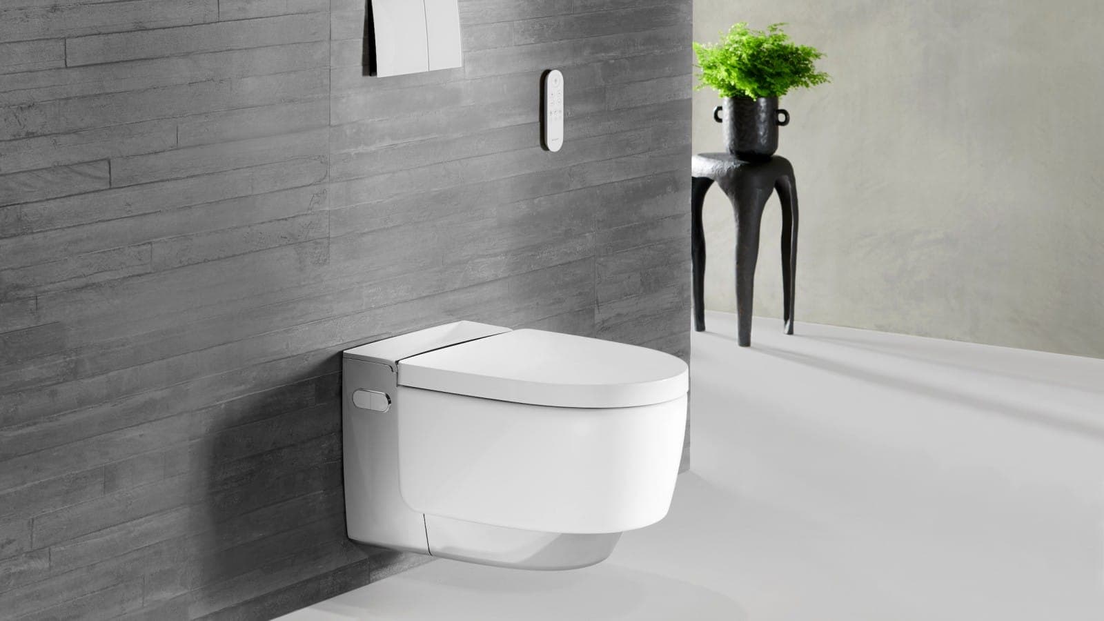 Inodoro con ducha AquaClean Mera blanco con detalle cromado, suspendido en un muro de losetas grises con textura de madera; arriba, placa actuadora blanca y control remoto; a la derecha, una mesa negra escultórica con una planta.