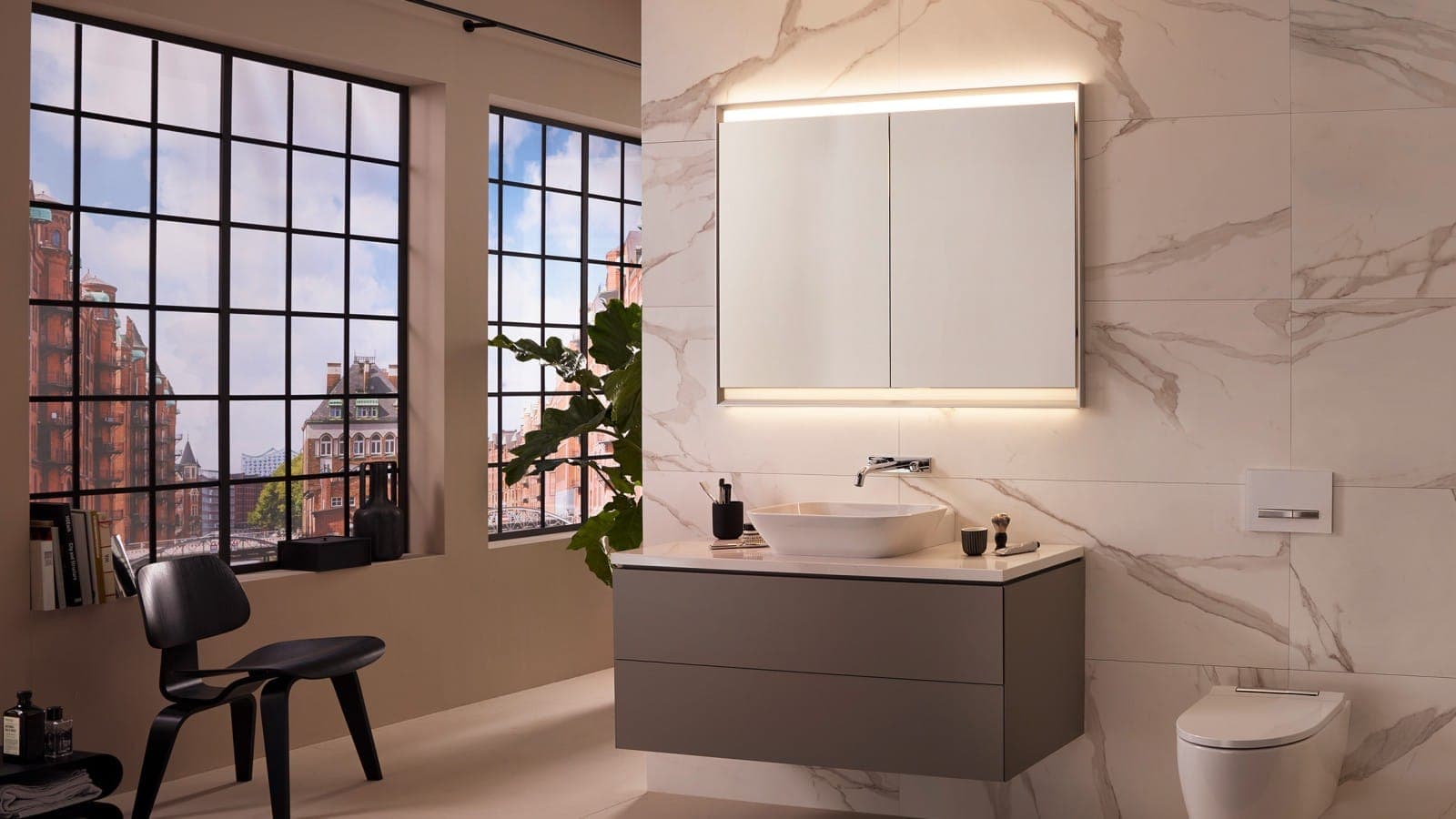 Baño con muros tipo mármol blanco: lavabo de sobreponer sobre mueble gris de dos cajones, grifo mural cromado y gabinete con espejo con luz cálida; a la derecha, un inodoro suspendido; a la izquierda, ventanales con vista a edificios de ladrillo y una silla negra.