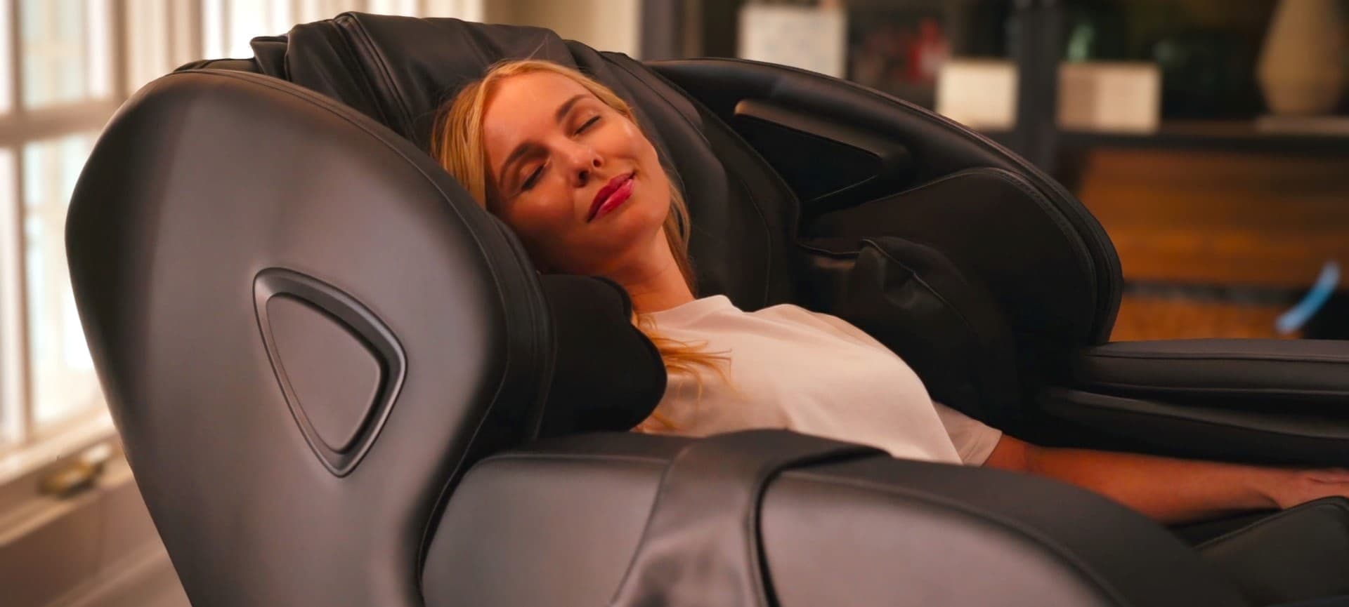 Mujer rubia con los ojos cerrados y una sonrisa, recostada en un sillón de masaje negro de respaldo envolvente; al fondo, una sala desenfocada con ventana y repisas.